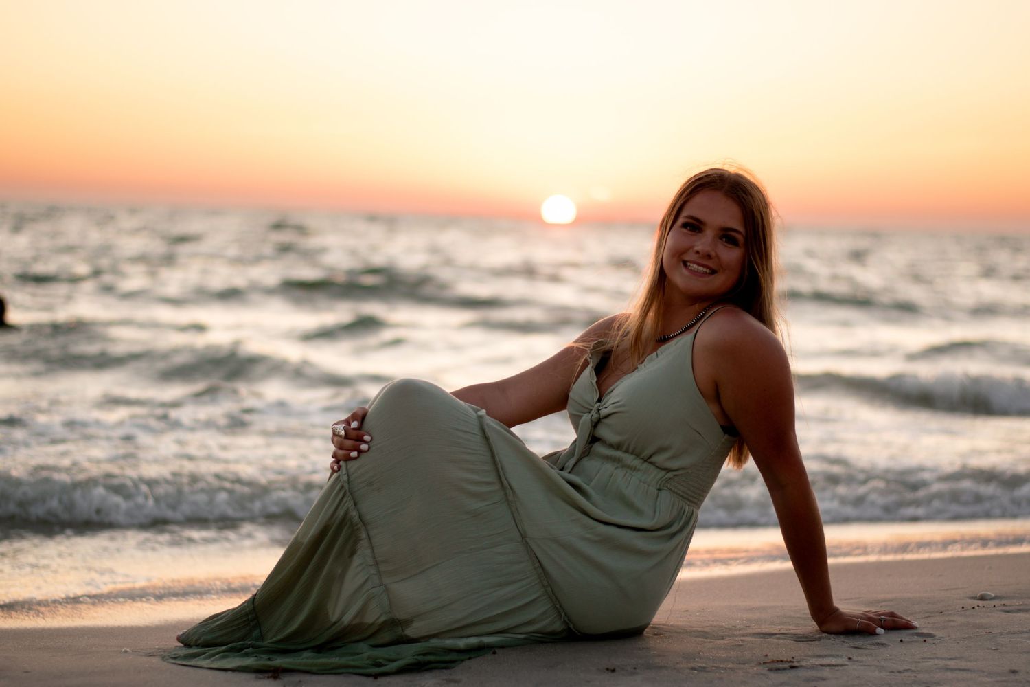 Smiling young woman in sage green maxi dress sitting on beach at golden sunset.