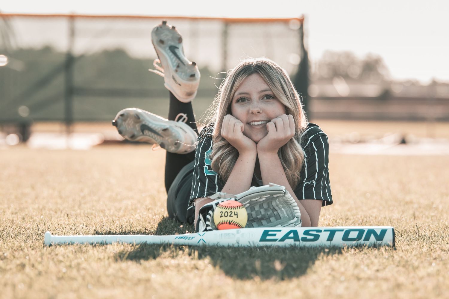Blonde softball player in uniform lies on grass field with Easton bat and cleats.