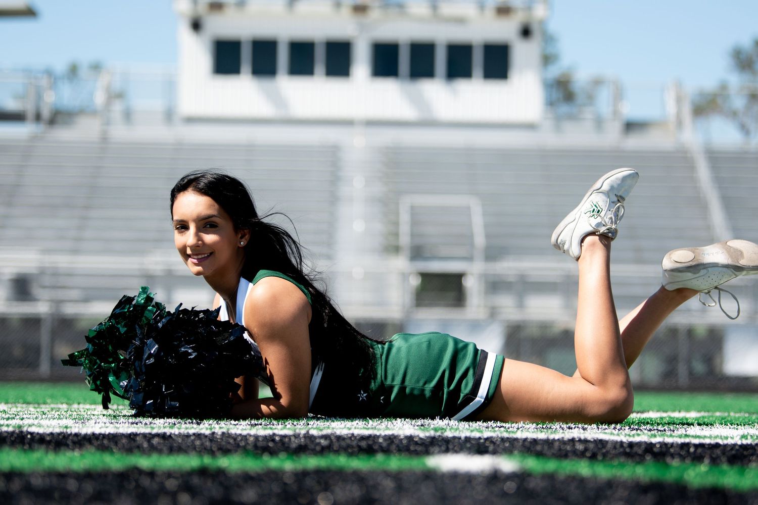 Cheerleader in green and white uniform posing with pom-poms on football field turf.