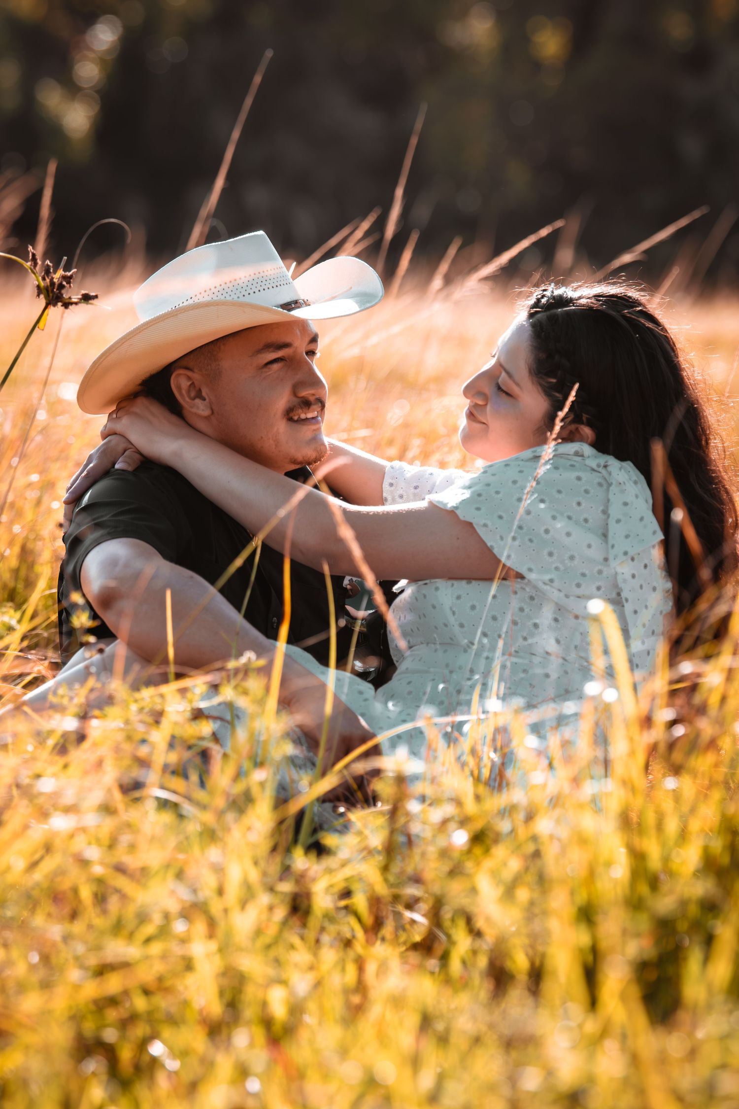 Romantic couple sitting together in golden sunlit field, woman embracing man in cowboy hat.