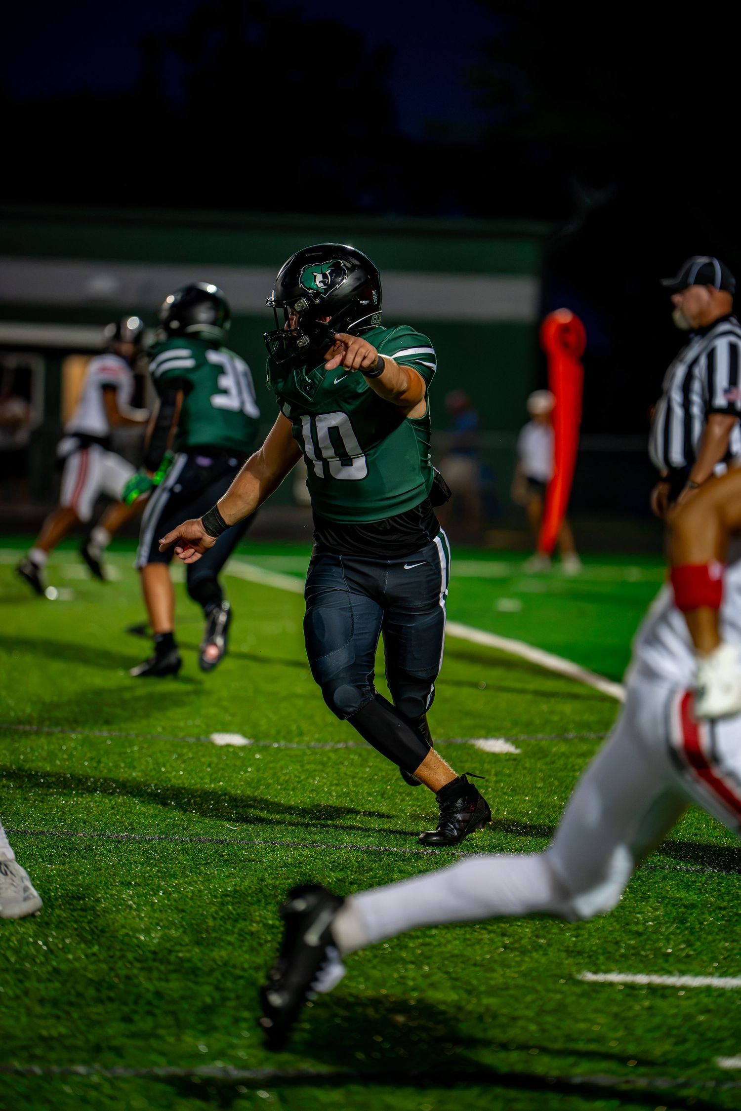 High school quarterback number 10 in green uniform scrambles during a night football game.
