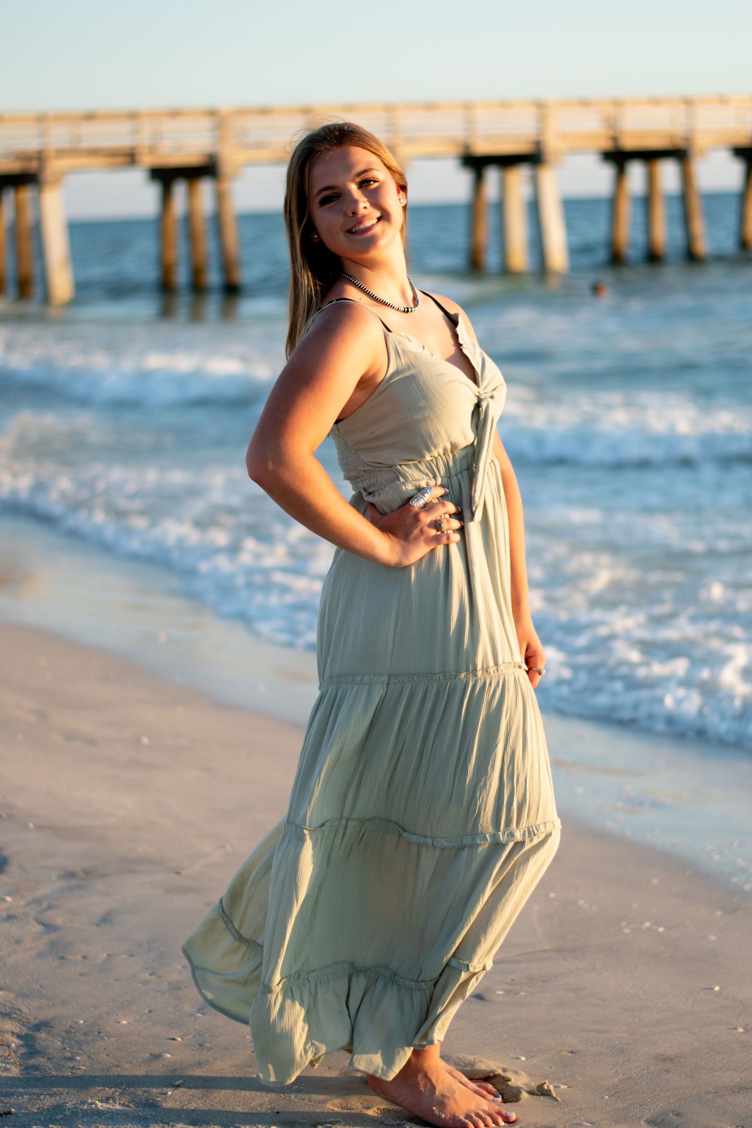 Smiling young woman in sage green maxi dress standing on beach near pier at sunset.