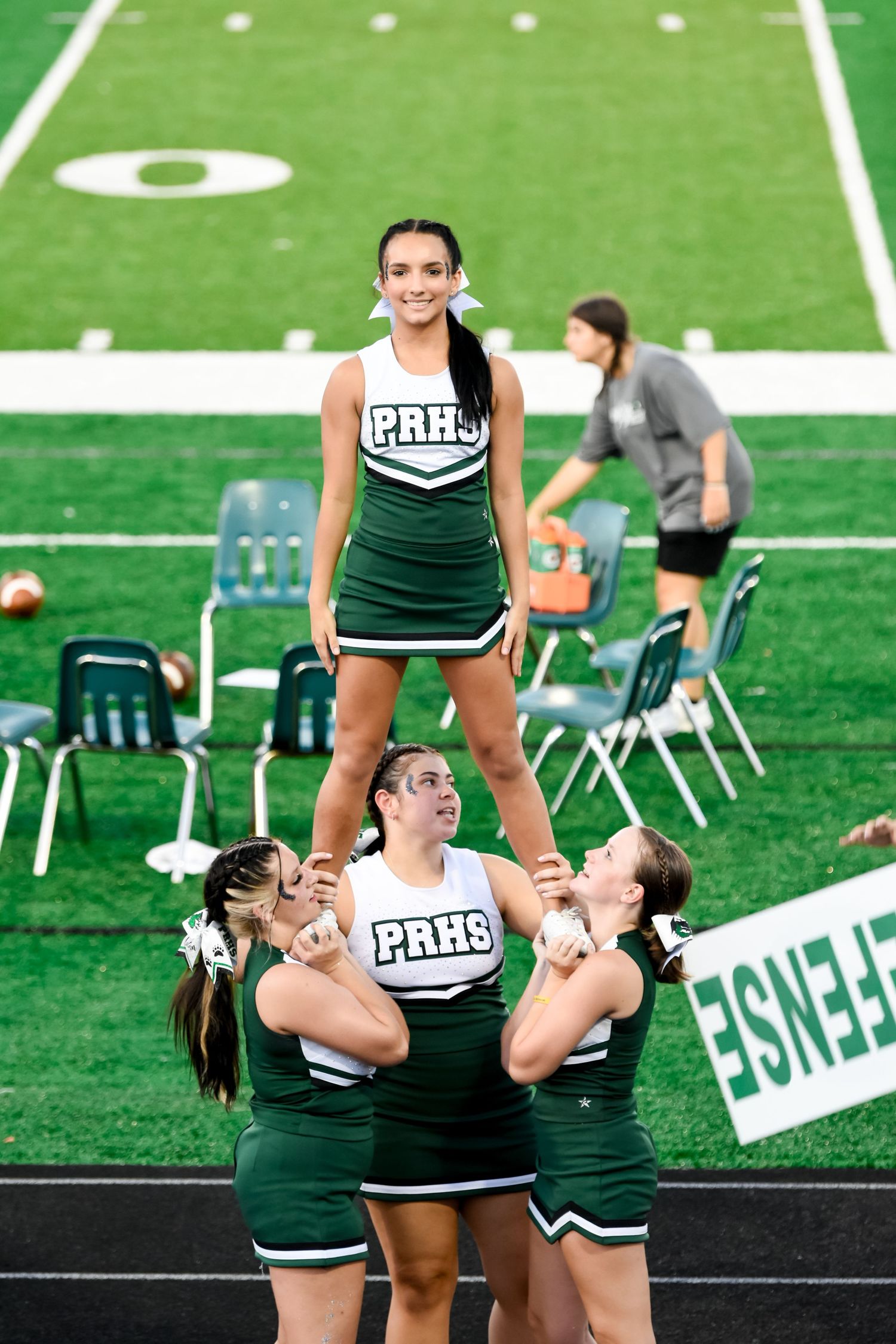 PRHS cheerleaders perform a stunt with a smiling flyer elevated above her bases on a football field sideline.