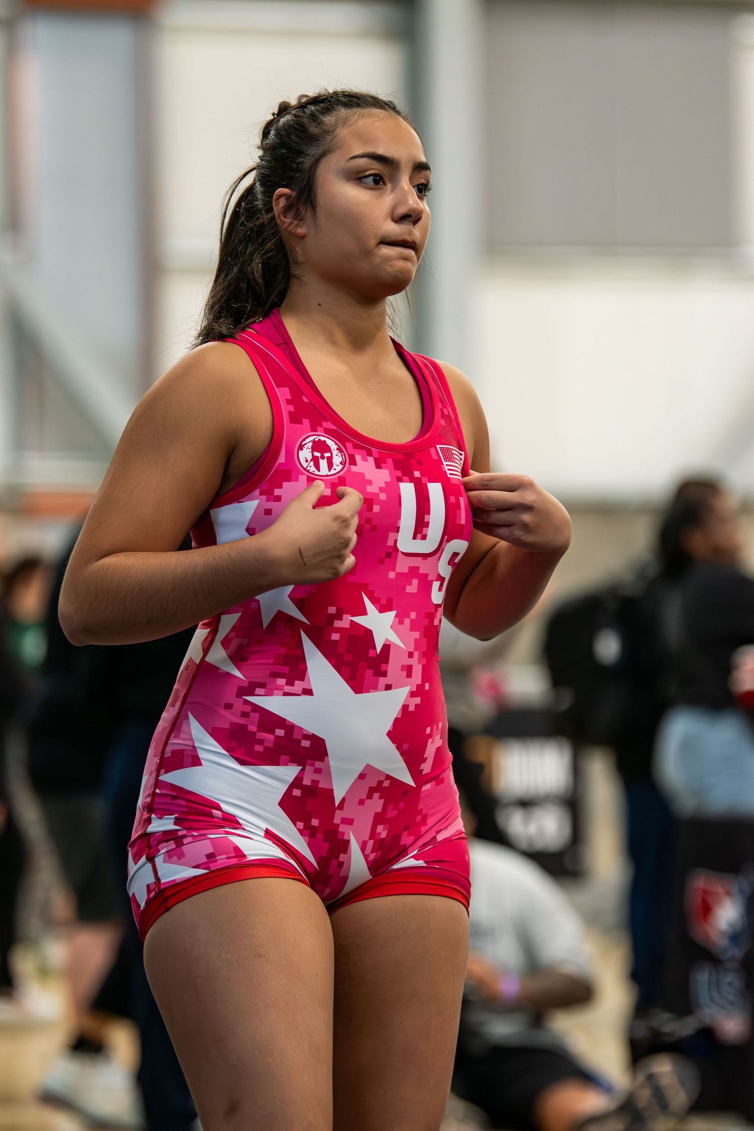 Young female wrestler in pink USA star-print singlet stands focused and ready before a wrestling match indoors.