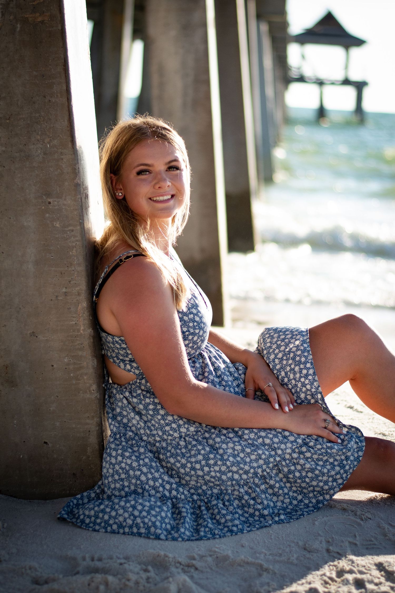 Smiling young woman in blue floral dress sitting under beach pier on sandy shore.