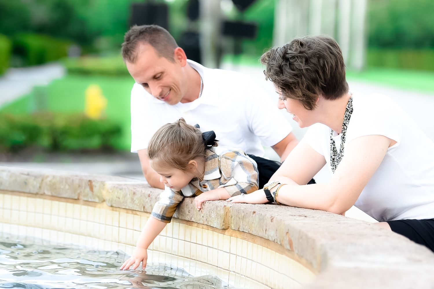 Family with young child leaning over decorative fountain in lush garden setting.