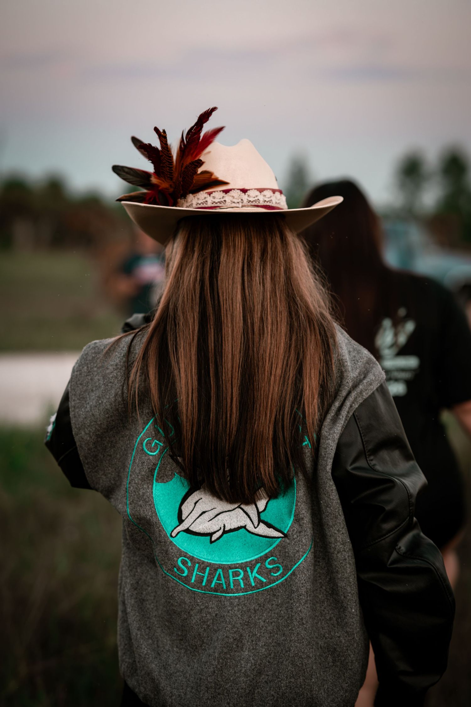 Back view of woman with long brown hair wearing feathered cowboy hat and Sharks jacket outdoors.