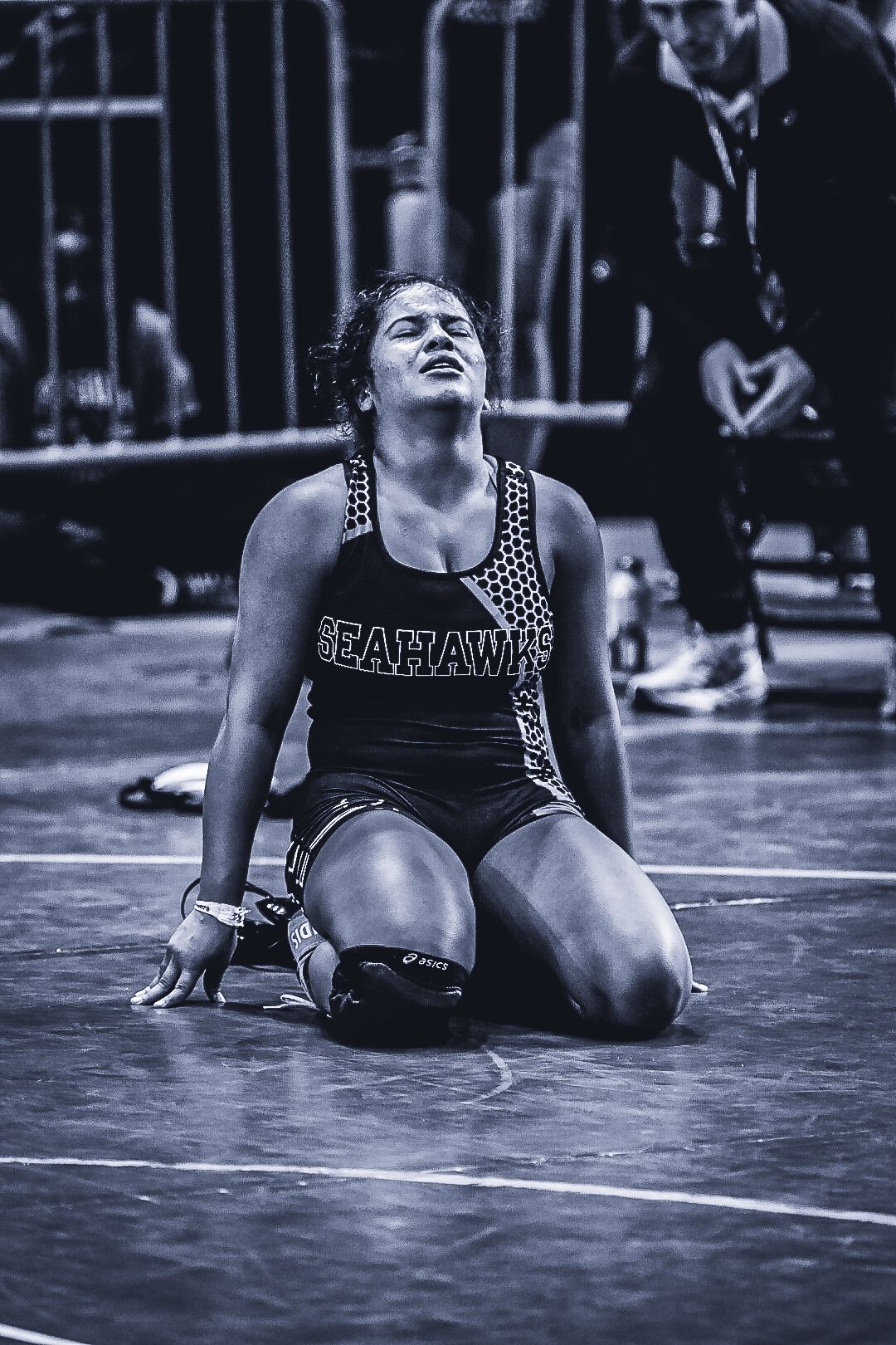 Emotional female Seahawks wrestler kneels on mat in black and white photo during competitive match.