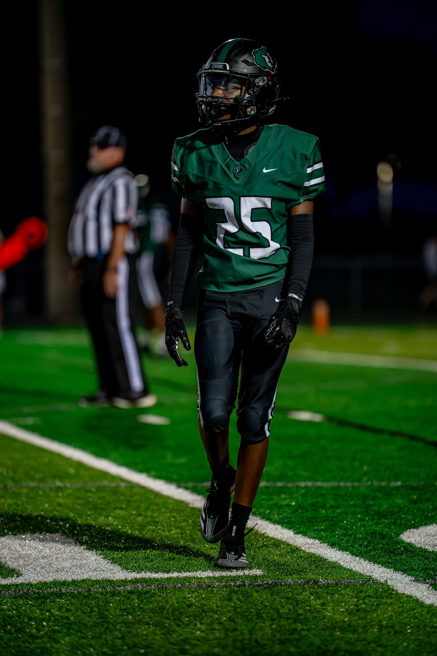 High school football player number 25 in green uniform walks on field during night game.