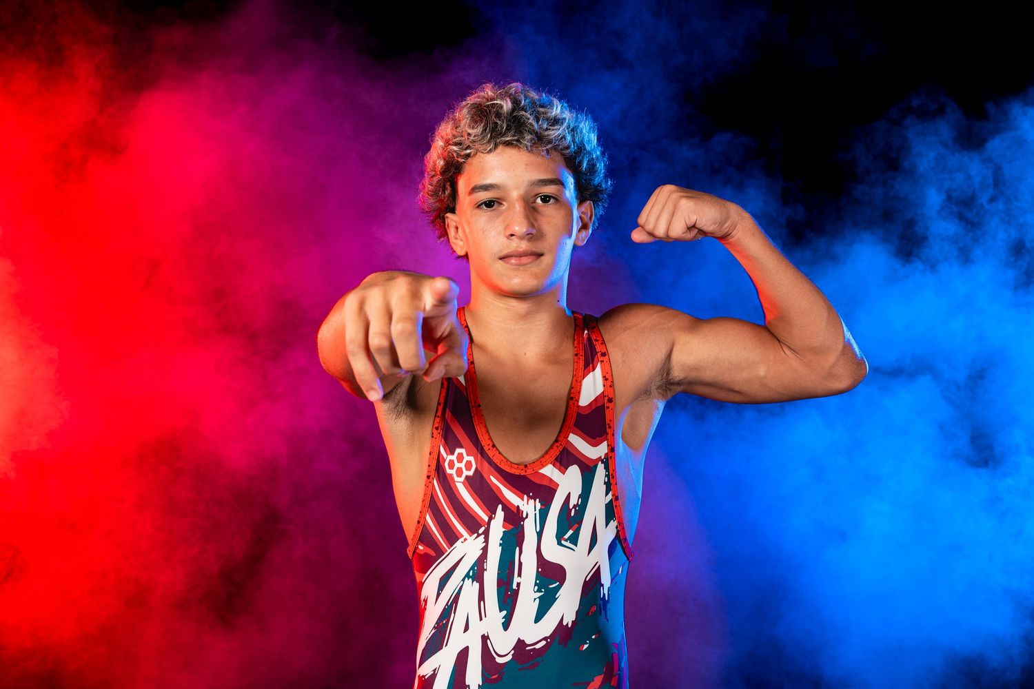 Wrestler in a USA singlet flexing and pointing toward the camera against red and blue smoke.