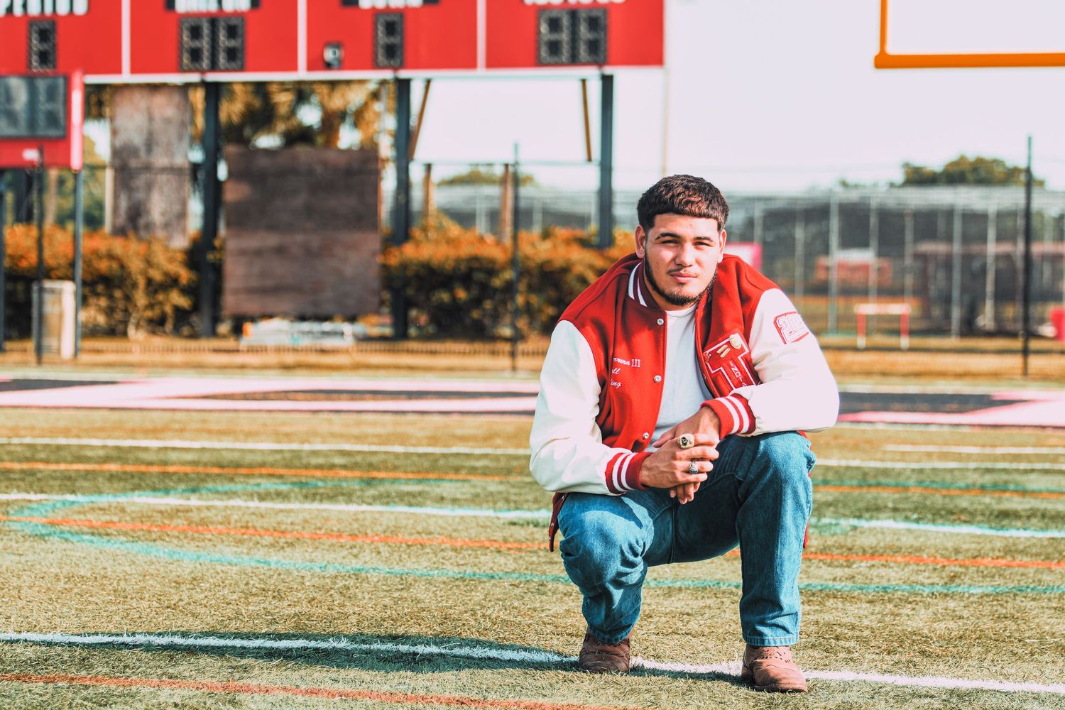 Young man in red letterman jacket crouching on high school football field turf.