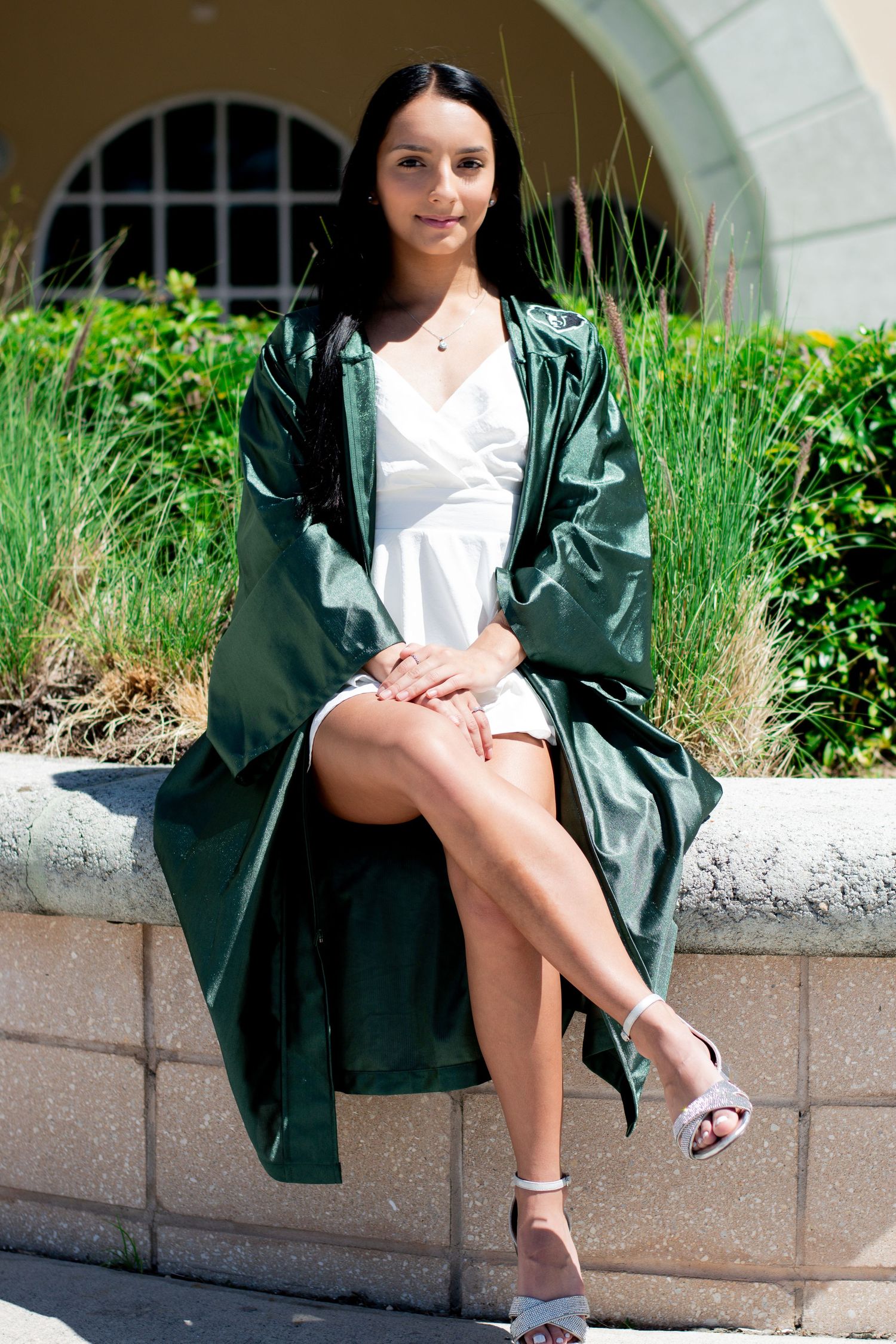 Young woman in green graduation gown and white dress sitting on stone wall outdoors.
