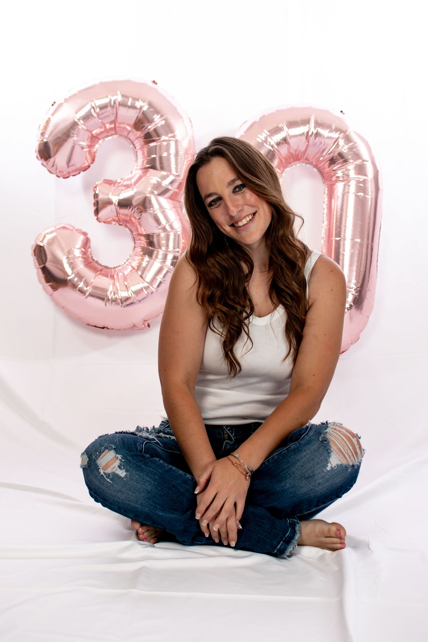 Happy woman sitting cross-legged in ripped jeans with pink rose gold number 30 balloons behind her.