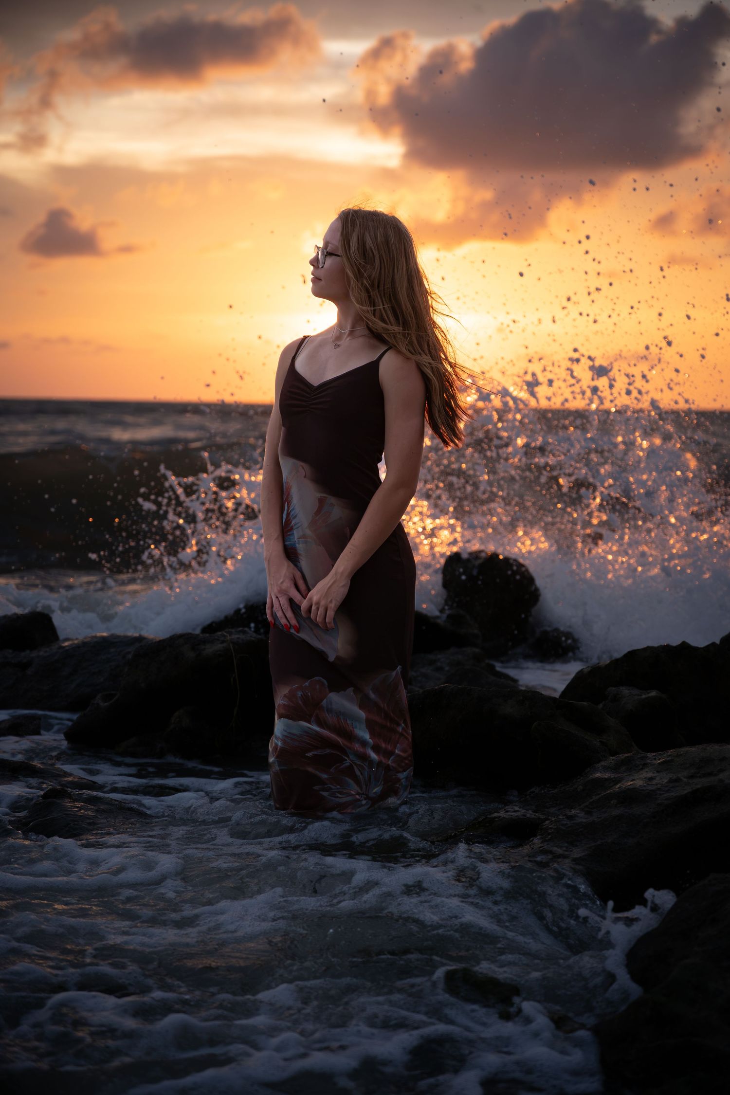Woman standing on rocky shore at sunset with dramatic ocean waves splashing behind her.