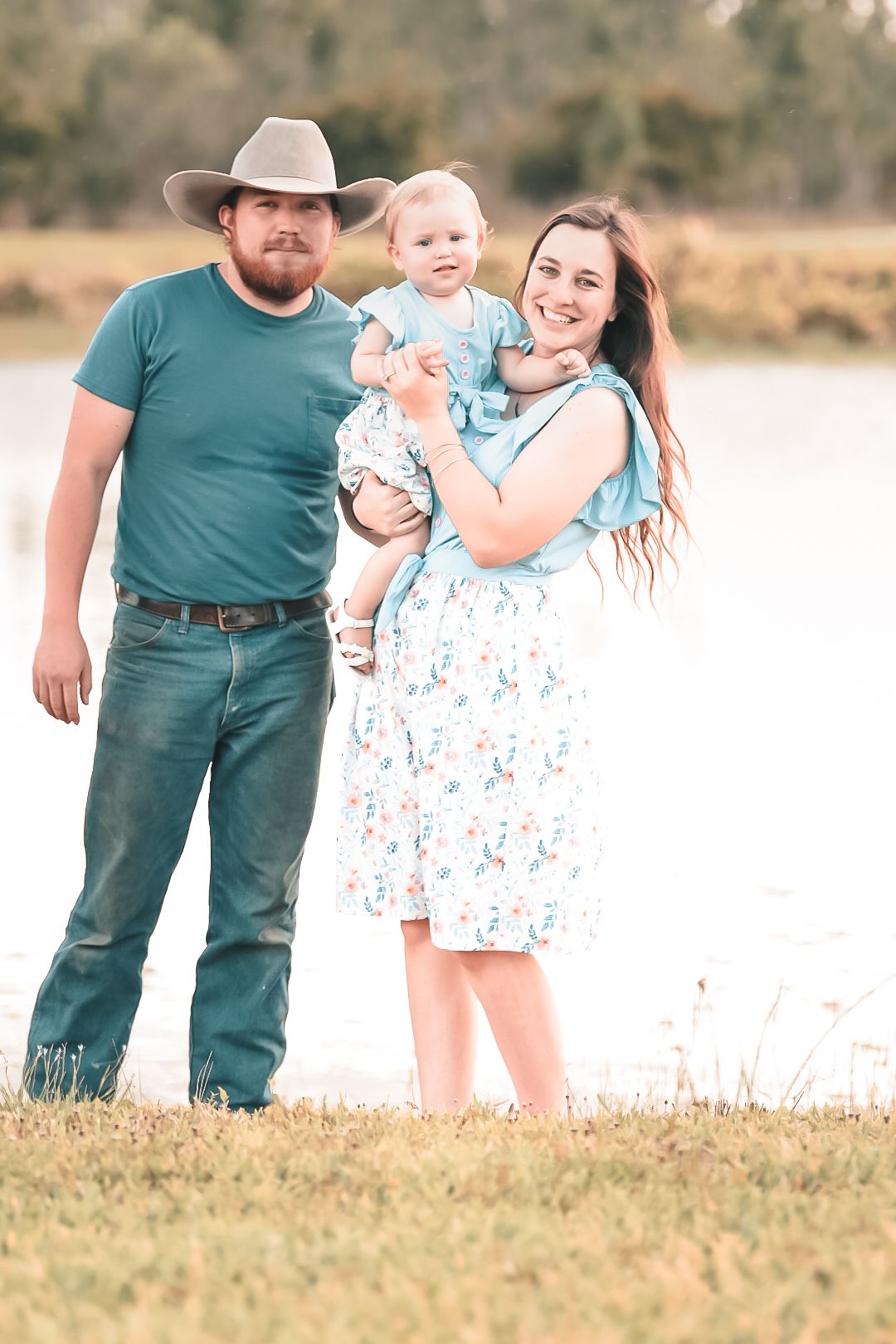 Young family with baby girl posing together near a peaceful lake in rural setting.