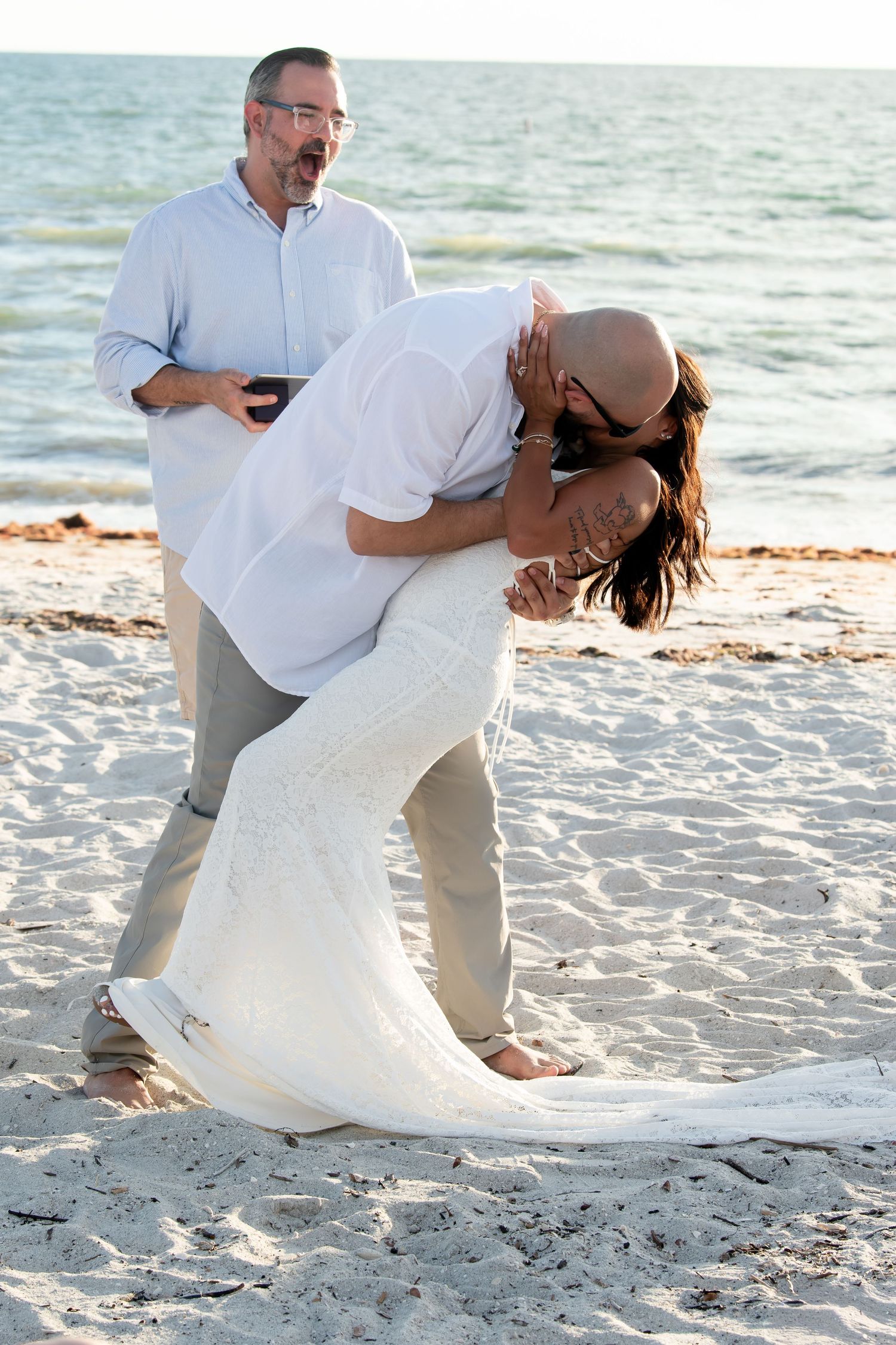 Groom dipping bride in white wedding dress for a kiss on sandy beach at sunset.