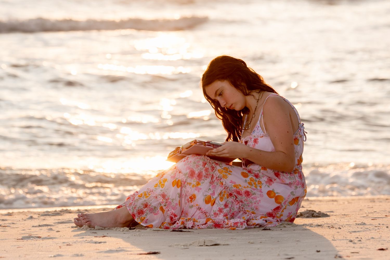 Young woman in floral dress sitting on sandy beach at golden sunset.