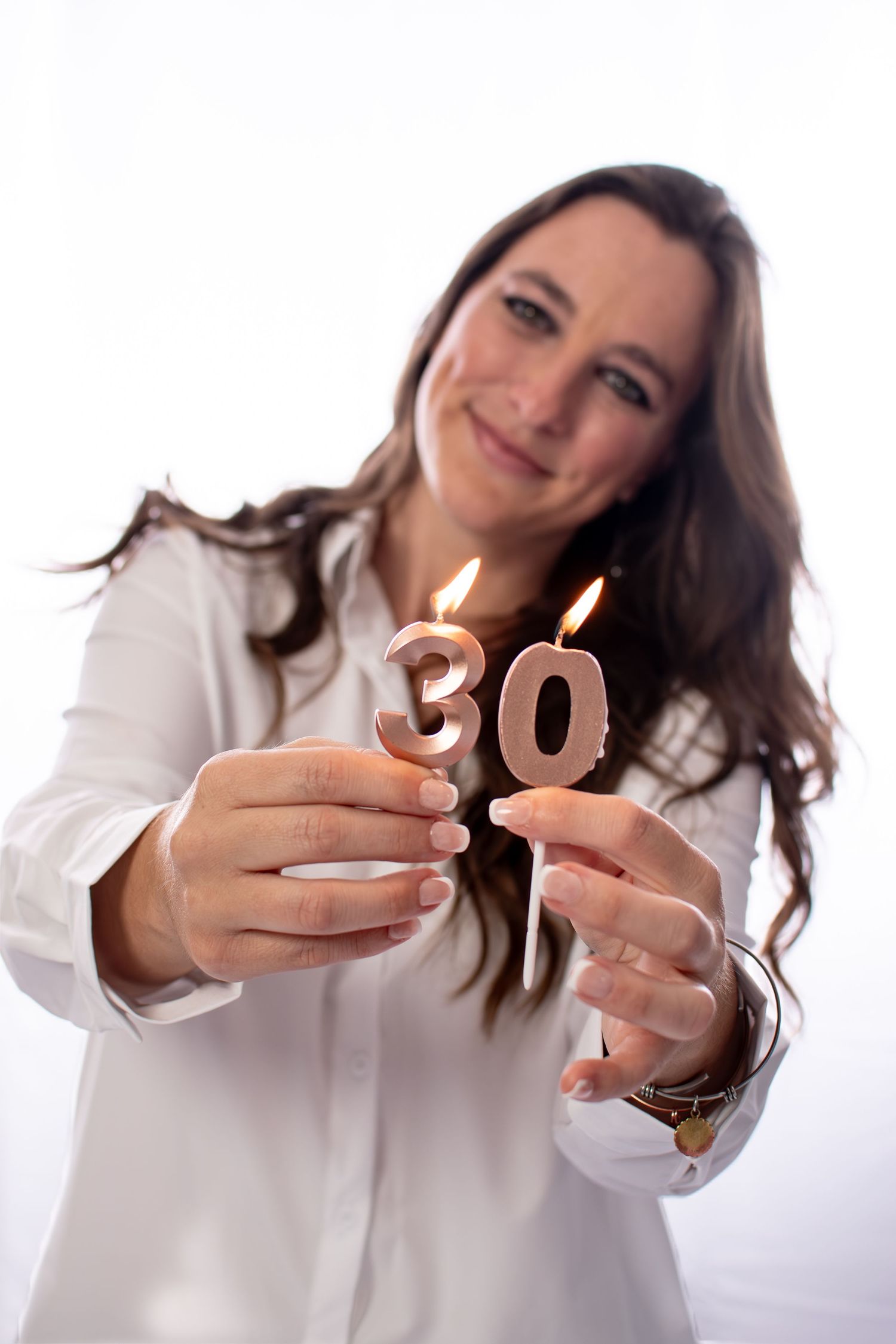 Woman in white shirt smiling while holding lit number 30 birthday candles on white background.