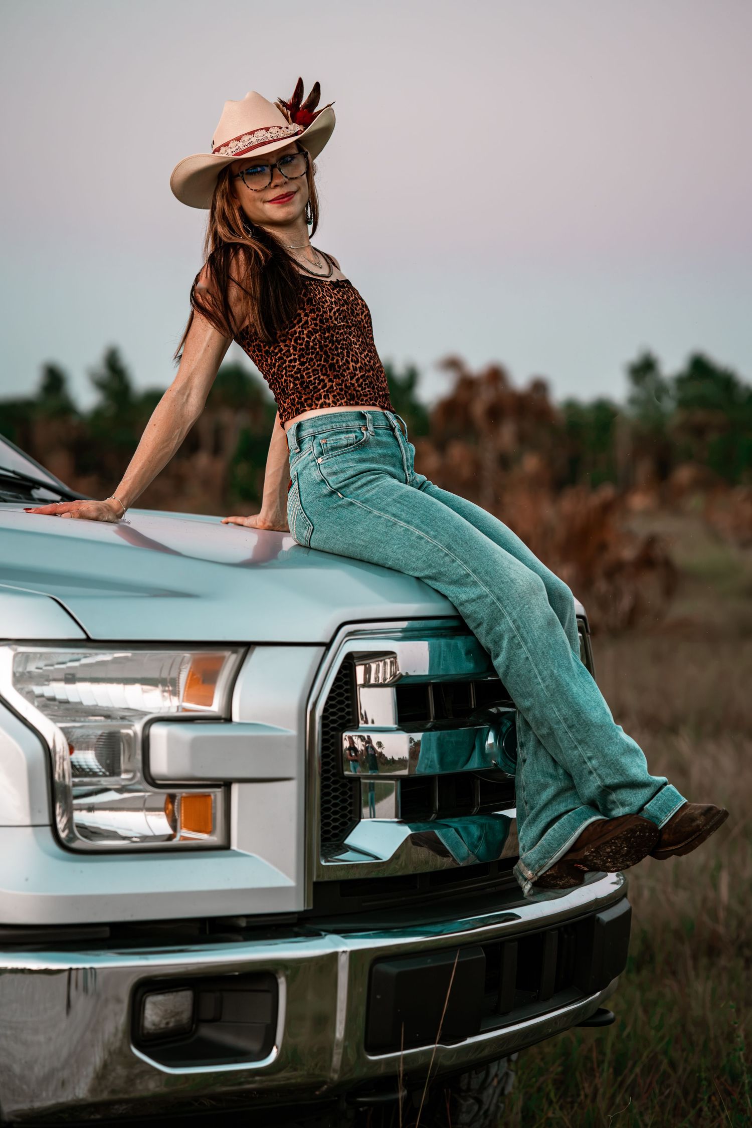 Woman in cowboy hat and leopard print top posing on hood of light blue Ford pickup truck.