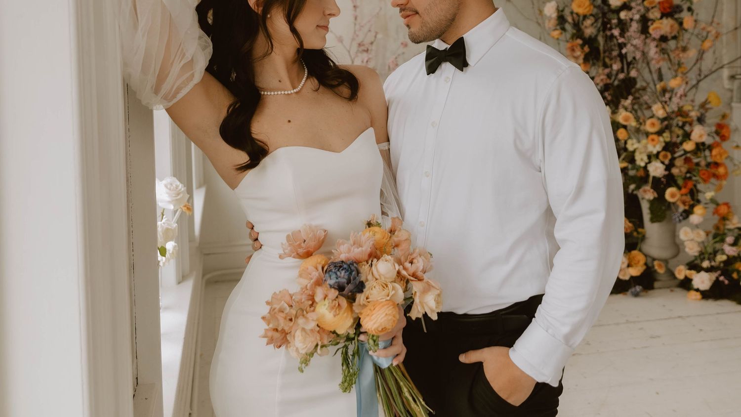 Wedding couple embraces and smiles together in front of floral backdrop with soft peach and cream flowers. luxury elopement ideas