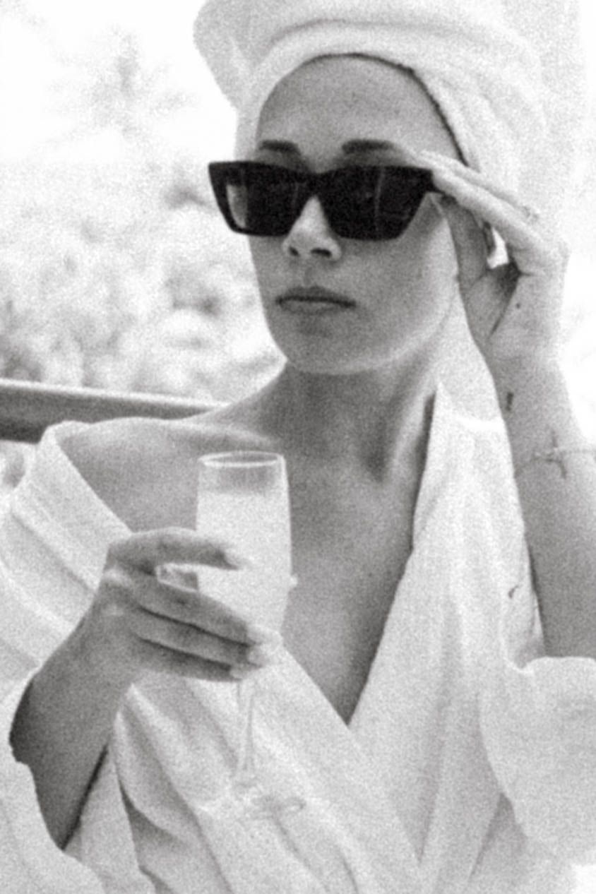 Glamorous retro-style portrait of woman in white robe and sunglasses holding beverage.