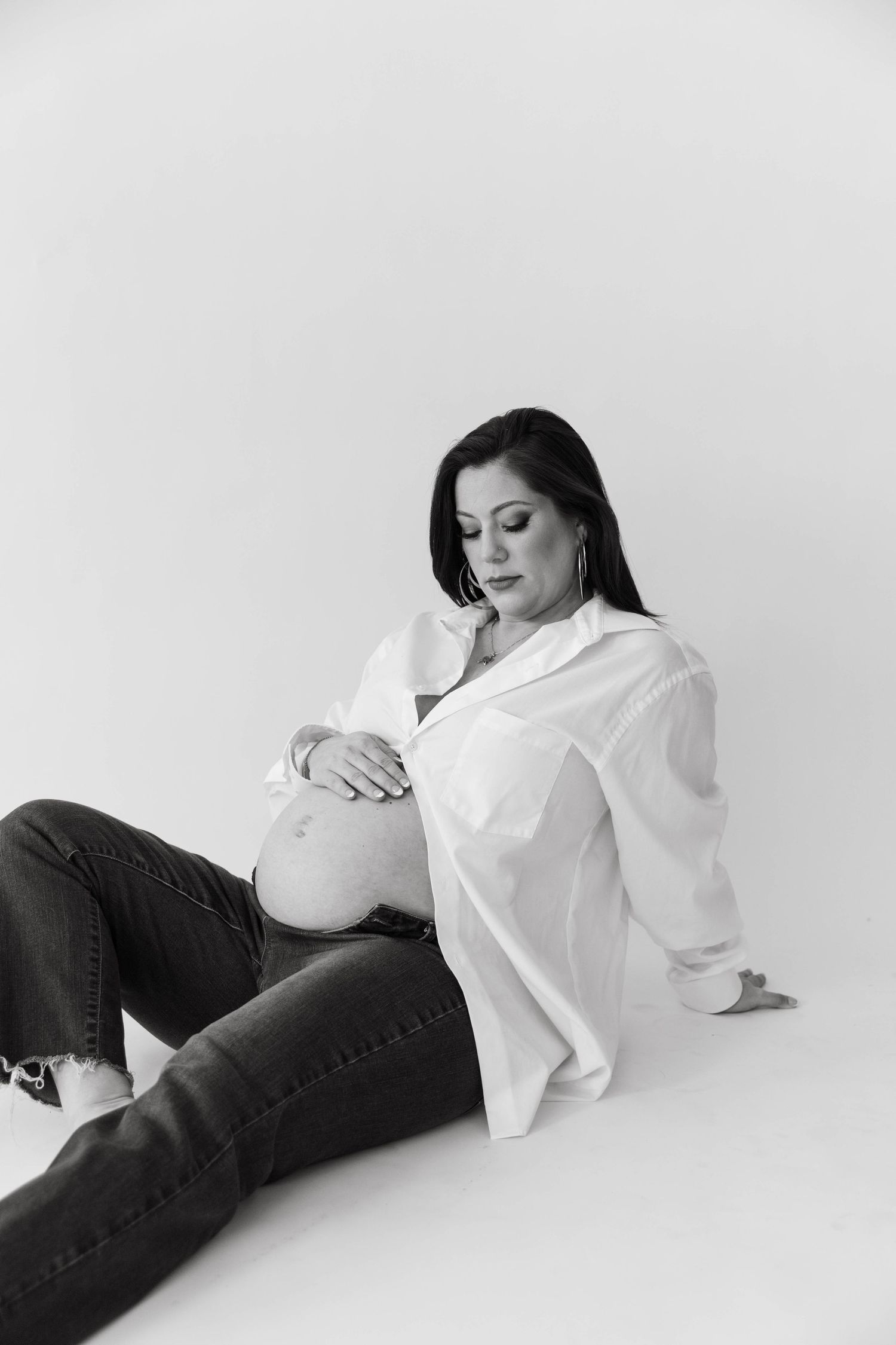 Maternity portrait in white shirt and jeans against minimalist background.