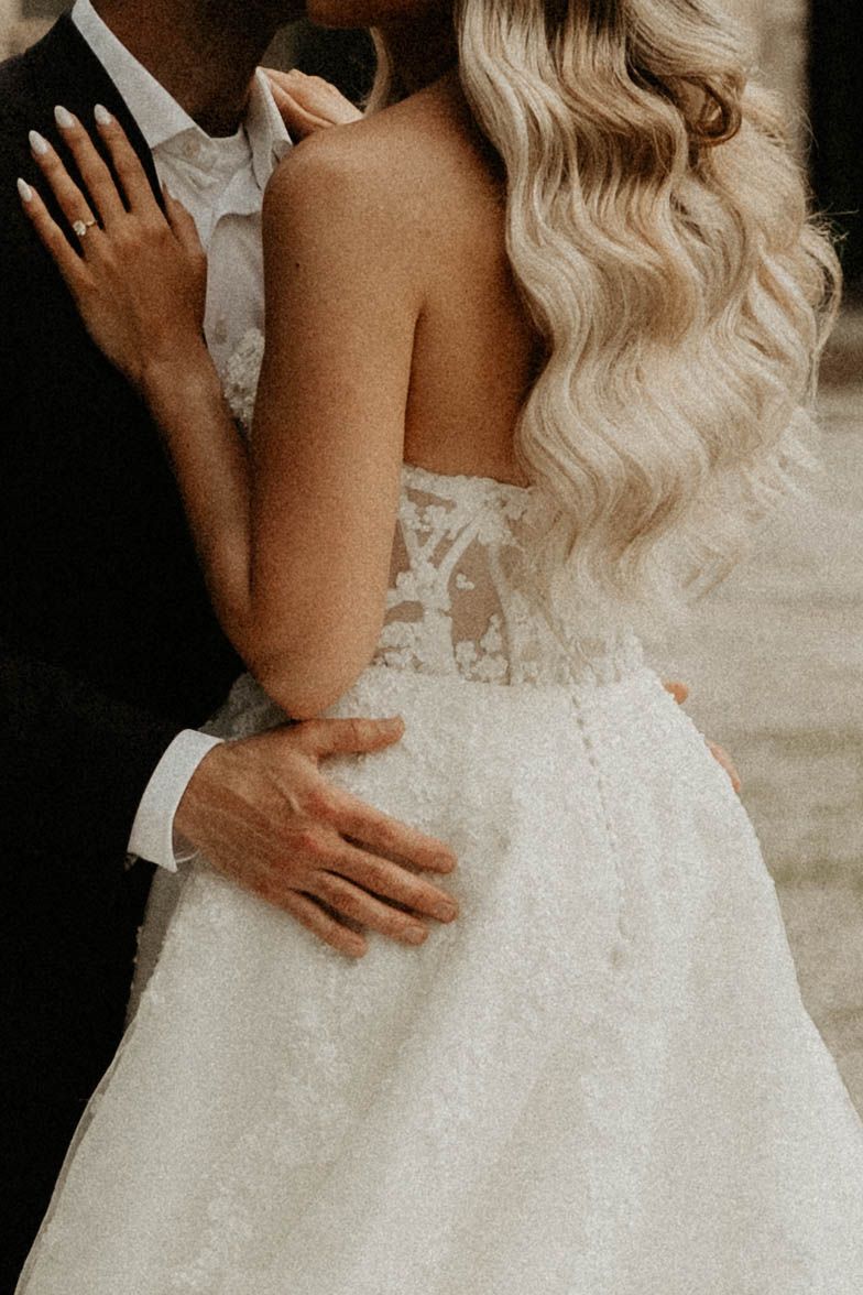 A romantic embrace showing the back of a wedding dress with flowing blonde waves and delicate lace details.