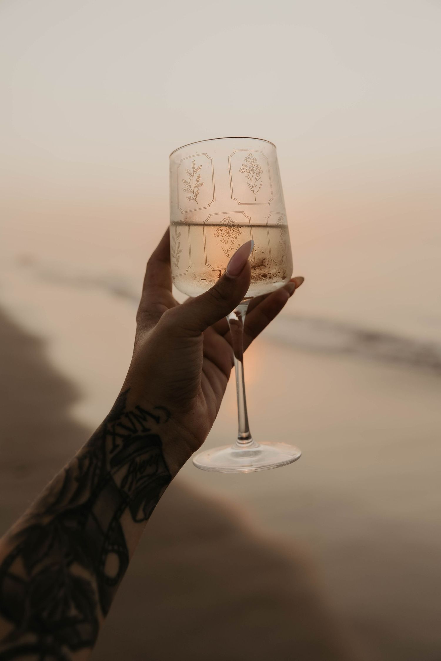 A tattooed hand holds up a wine glass with etched leaf designs against a hazy sunset background.