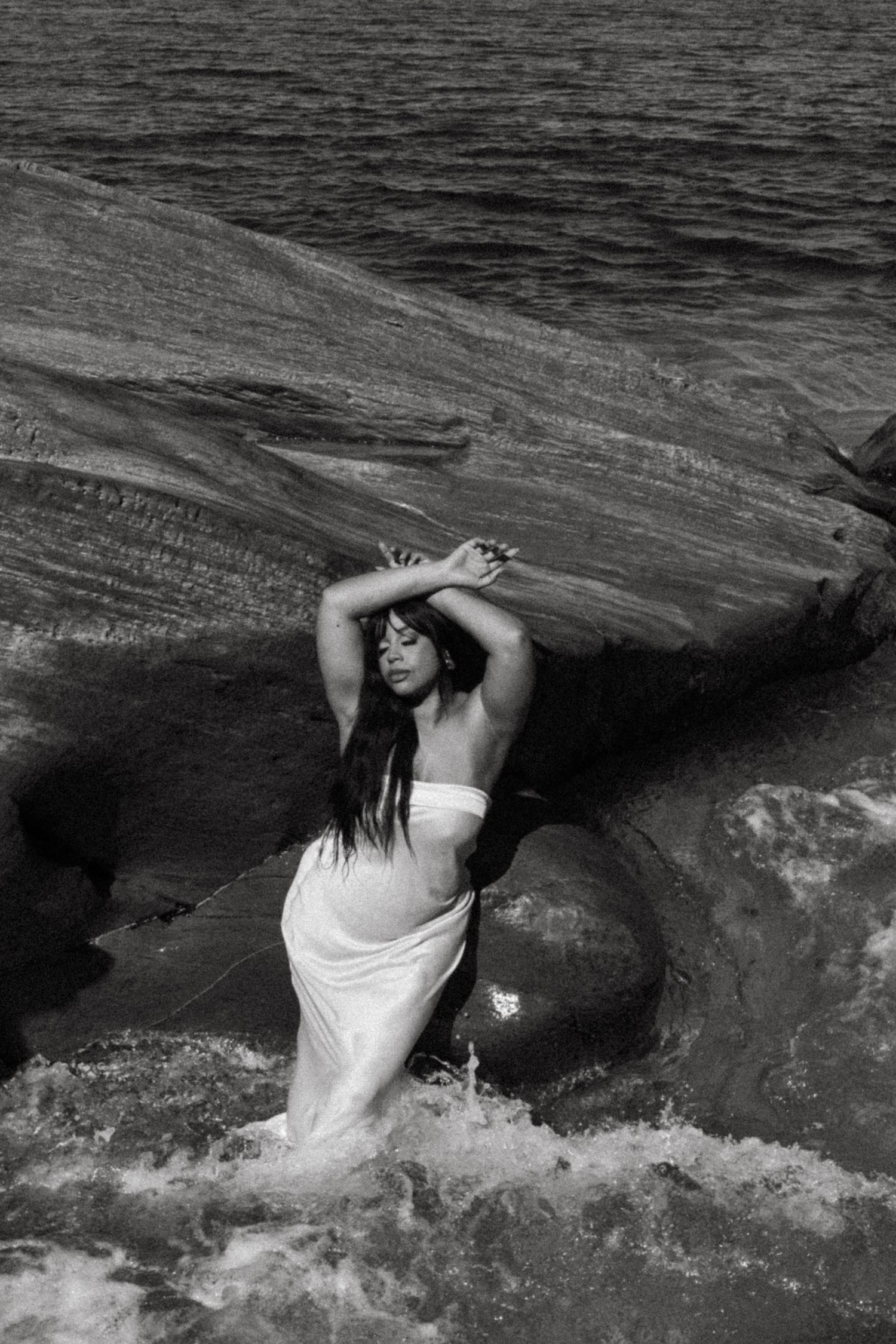 Dramatic black and white photo of figure in white dress posed among ocean rocks with crashing waves.
