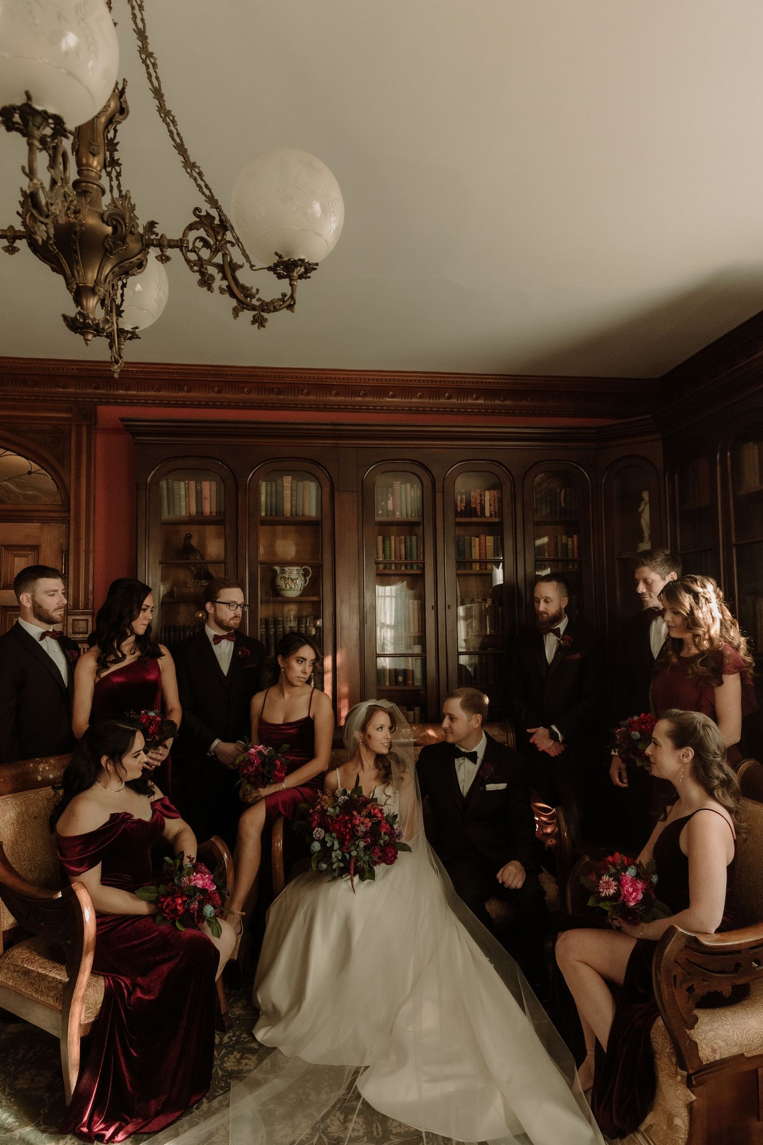 Wedding party posed in an elegant library with burgundy bridesmaids dresses and formal attire.
