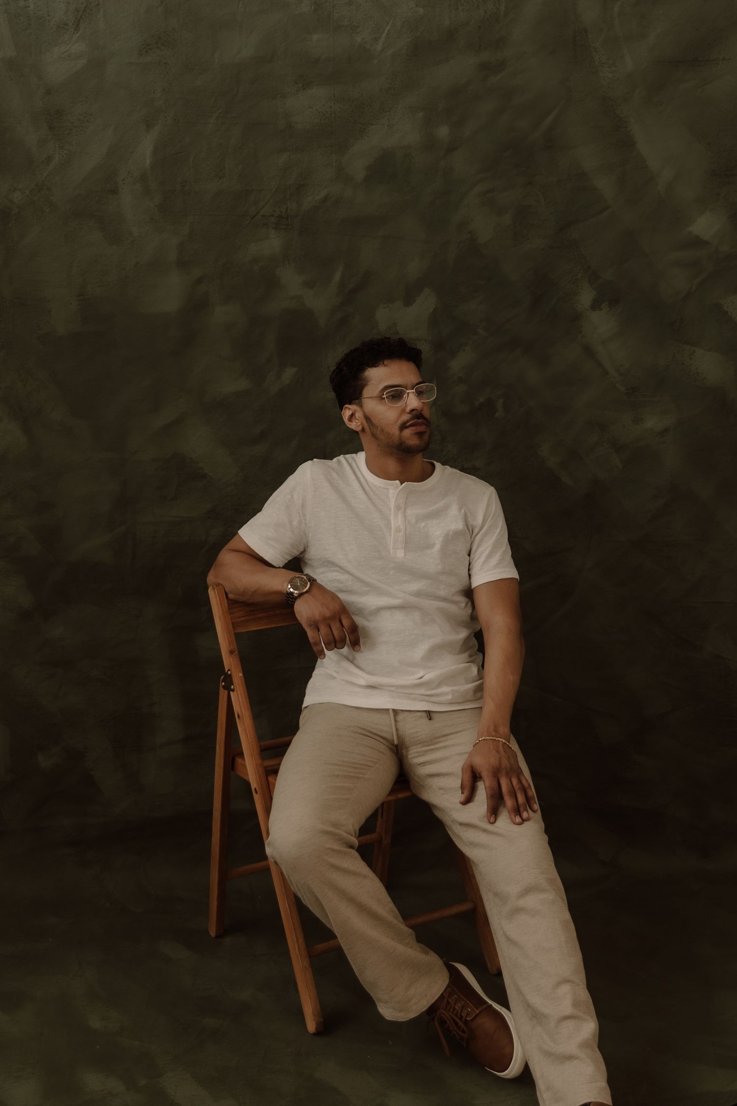 A person in a white henley shirt and beige pants sits casually on a wooden chair against a dark textured background.