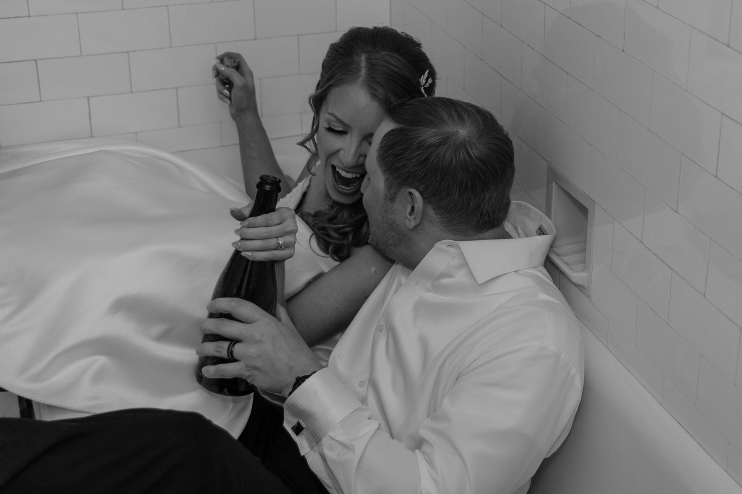 A couple shares an intimate black and white moment laughing together while sitting in a bathtub fully clothed.