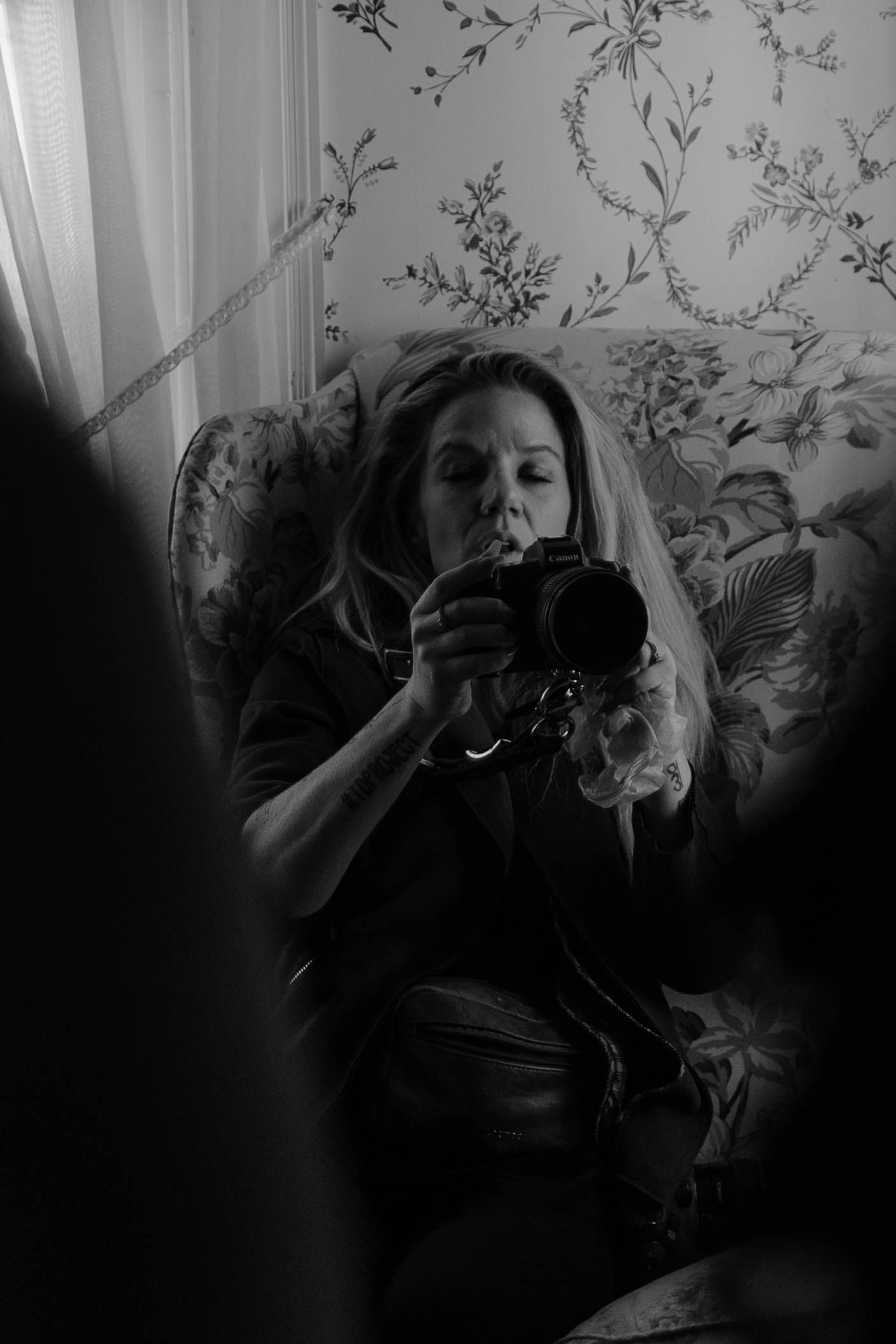 Black and white self portrait of a photographer reflected in a mirror with floral wallpaper.