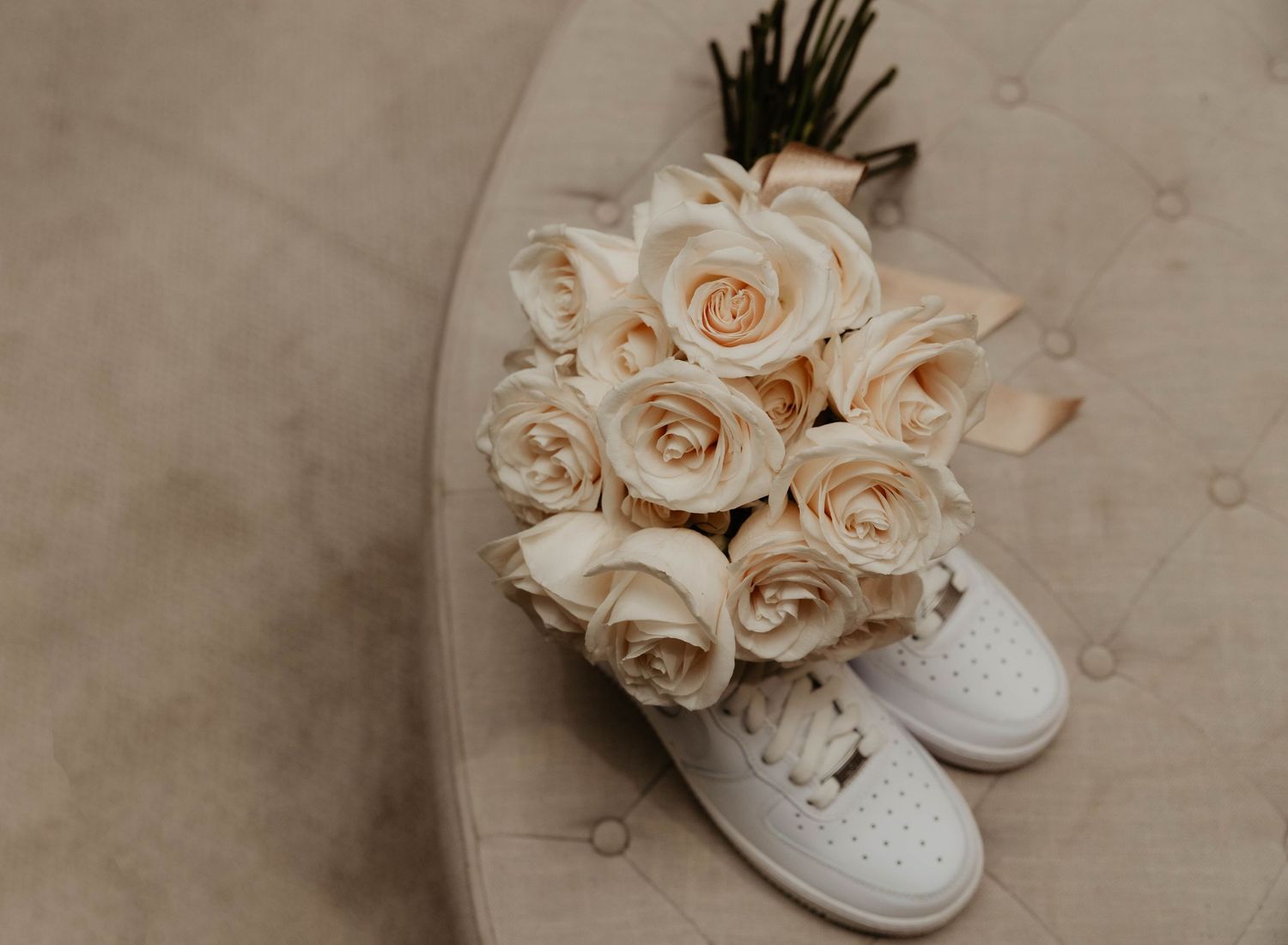 A delicate bouquet of cream-colored roses rests atop white sneakers on a beige textured surface.