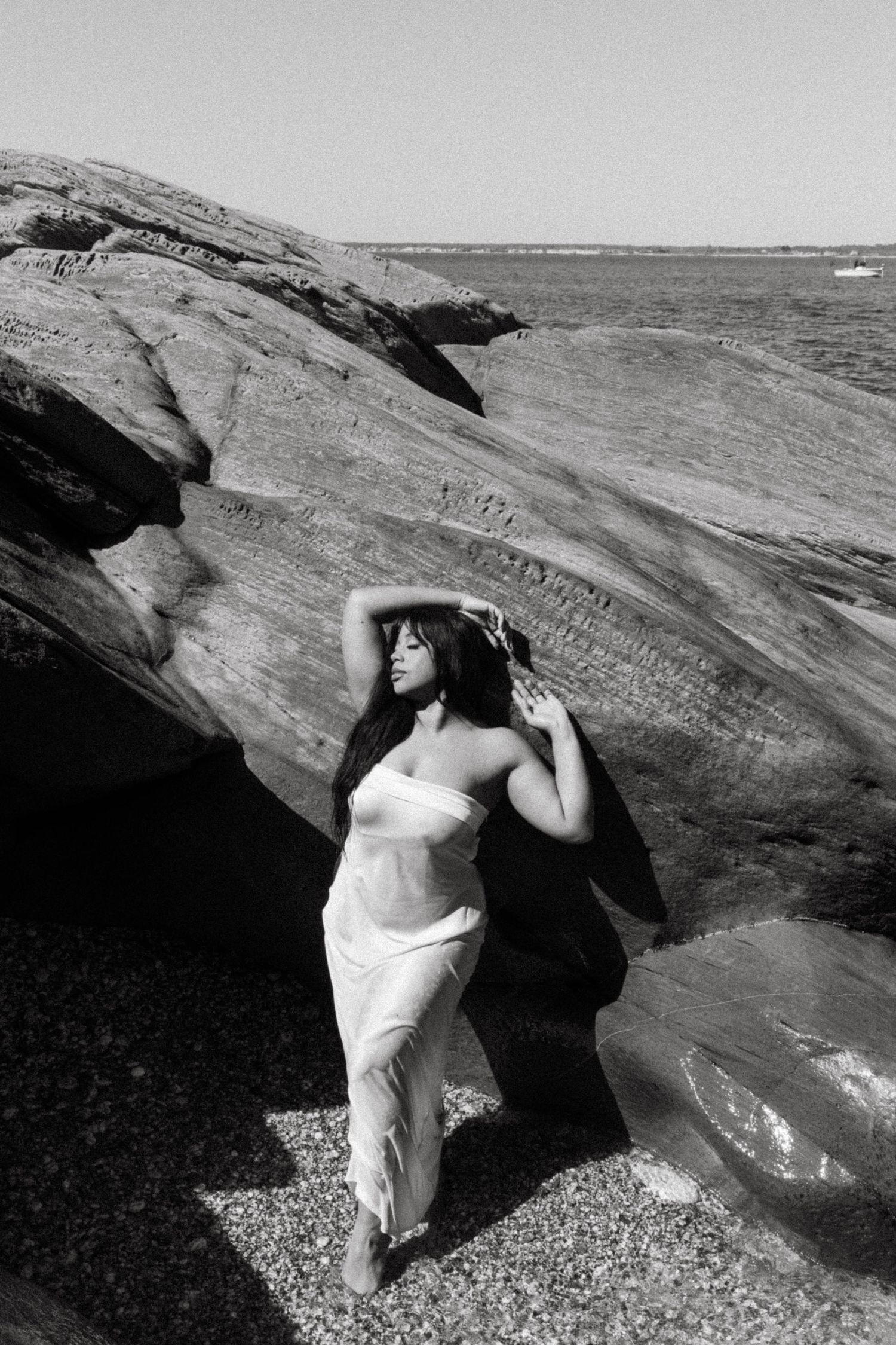Artistic black and white photo of figure in flowing white dress posed against rocky coastal backdrop.