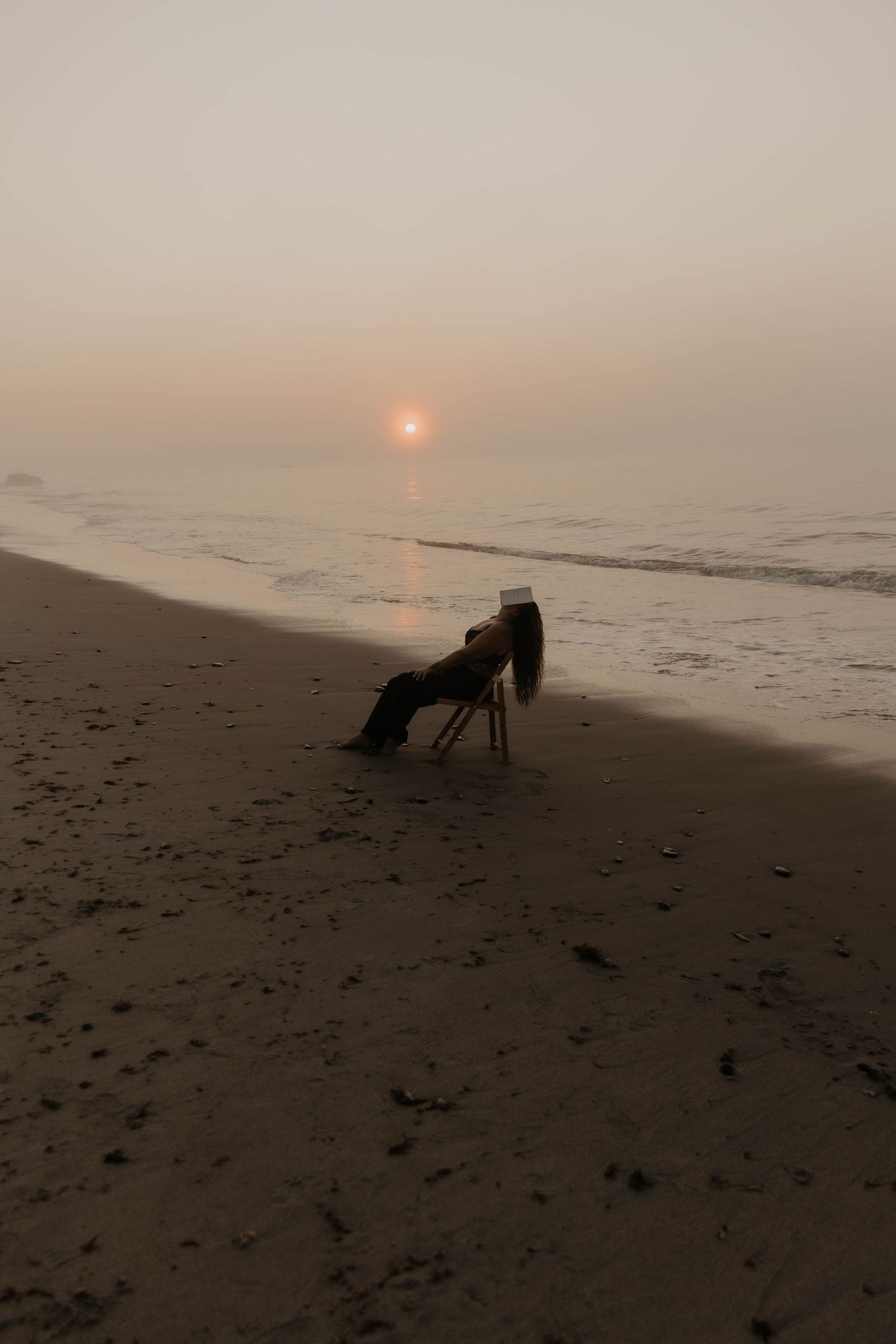beach chair silhouetted against a hazy sunset over the ocean with waves rolling onto the sandy shore.