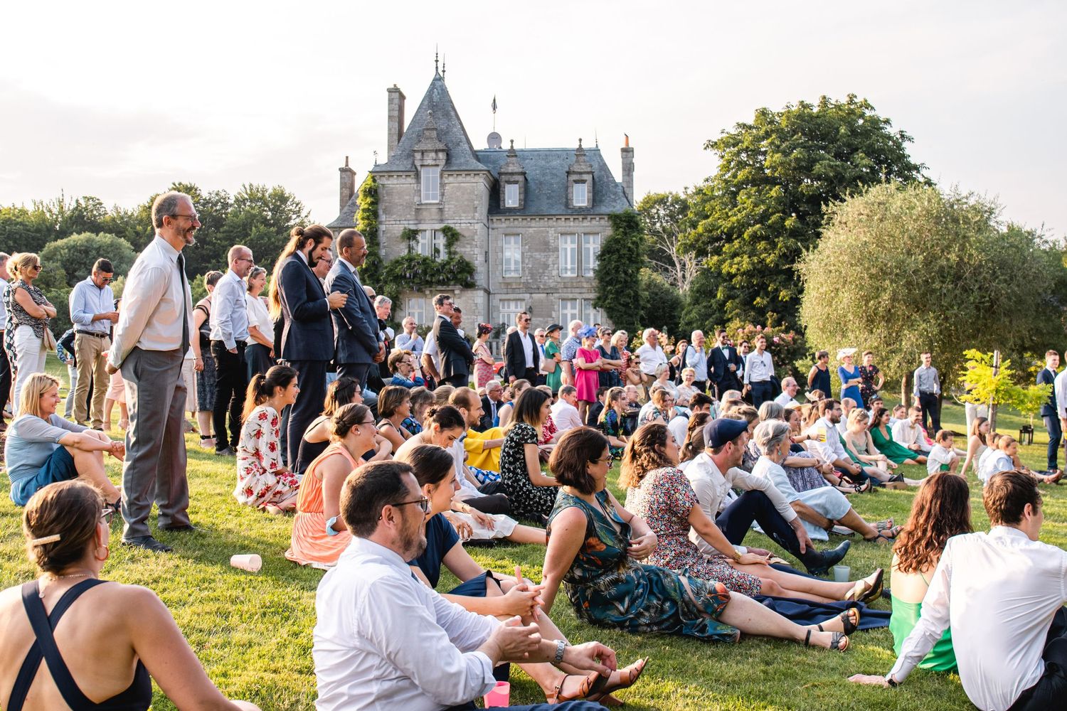 Wedding guests gather outdoors on a grassy lawn near a scenic chateau.
