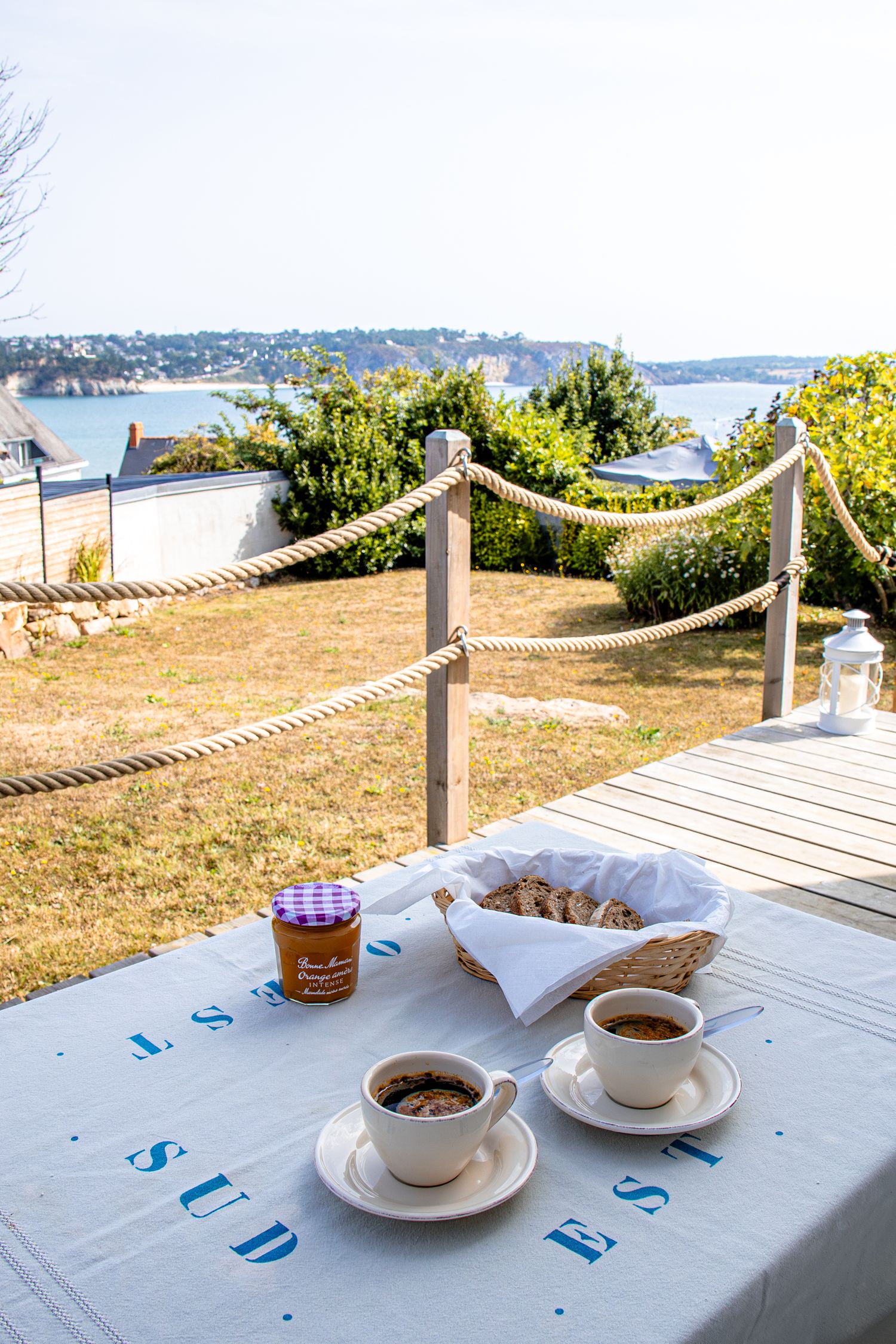 Coffee cups and pastries on a white tablecloth overlooking a scenic waterfront view on a sunny day.