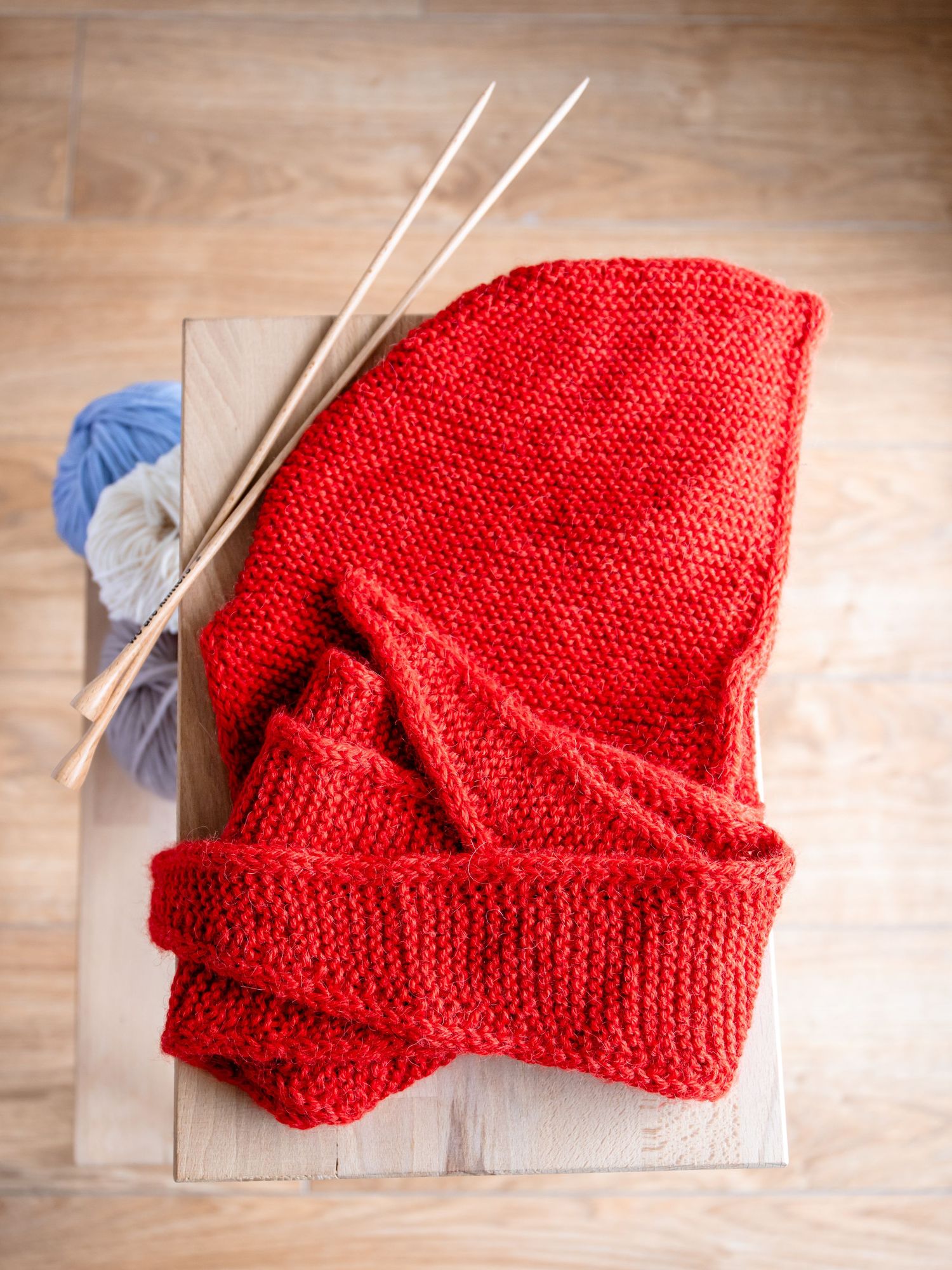 Bright red knitted fabric with knitting needles and blue yarn balls on wooden background.