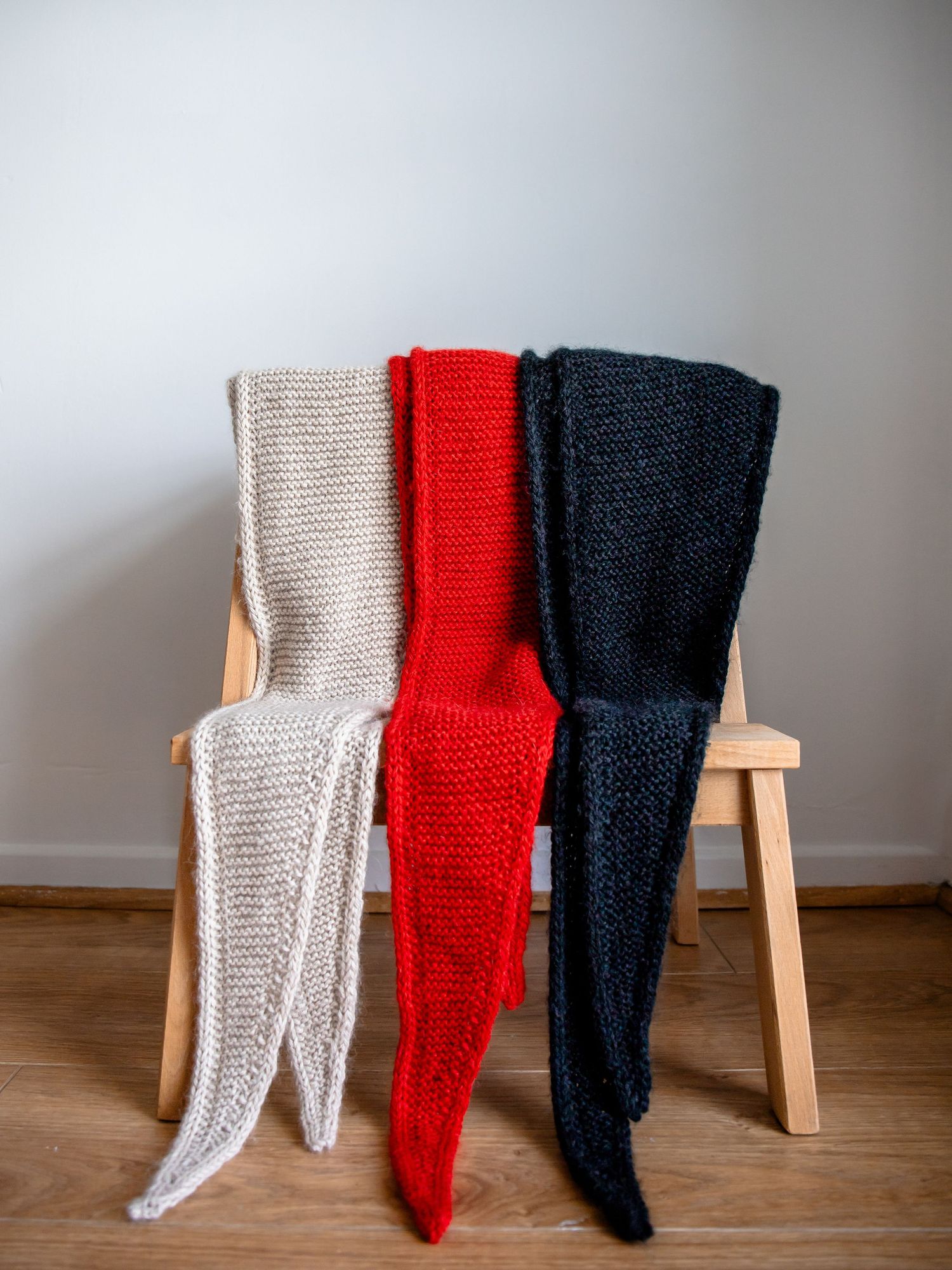 Three knitted scarves in beige, red, and navy blue hanging on a wooden chair against a white wall.