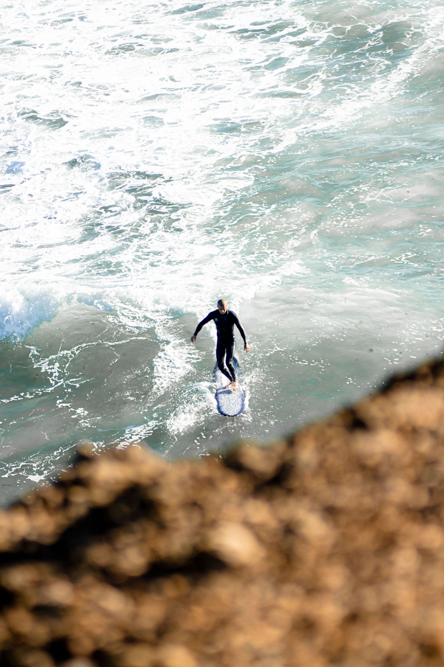 Surfer riding a wave along rocky coastline viewed from above.