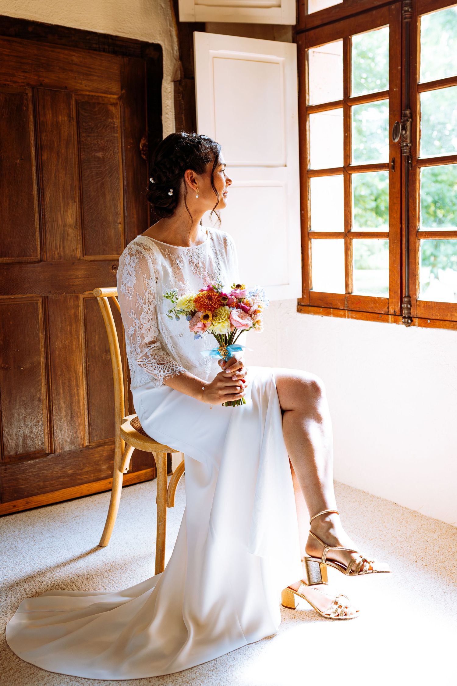 A bride in a long-sleeved lace wedding dress sits by a window holding a colorful bouquet in a rustic room.