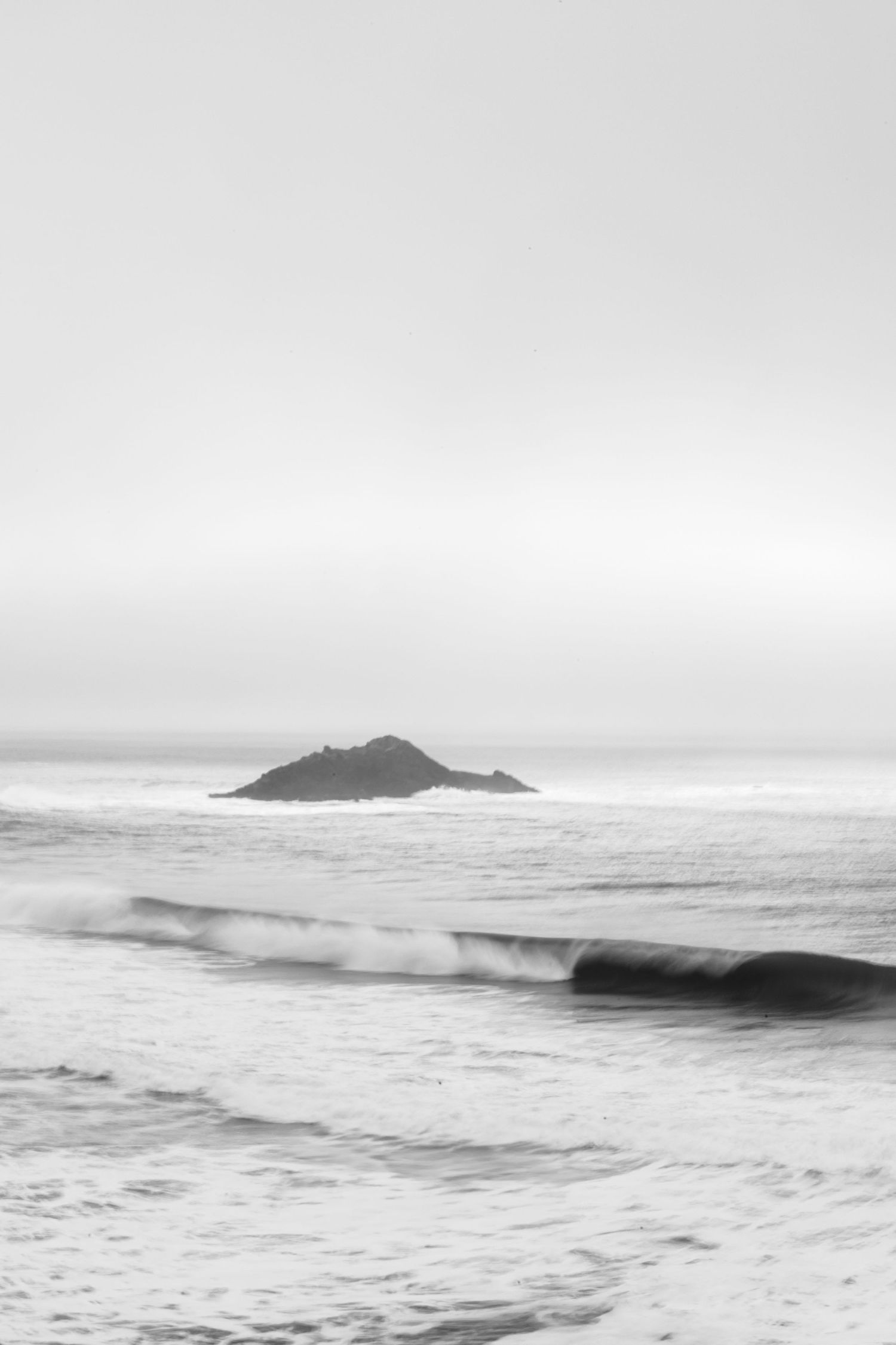 A black and white seascape featuring rolling waves and a small rocky island emerging from misty waters.