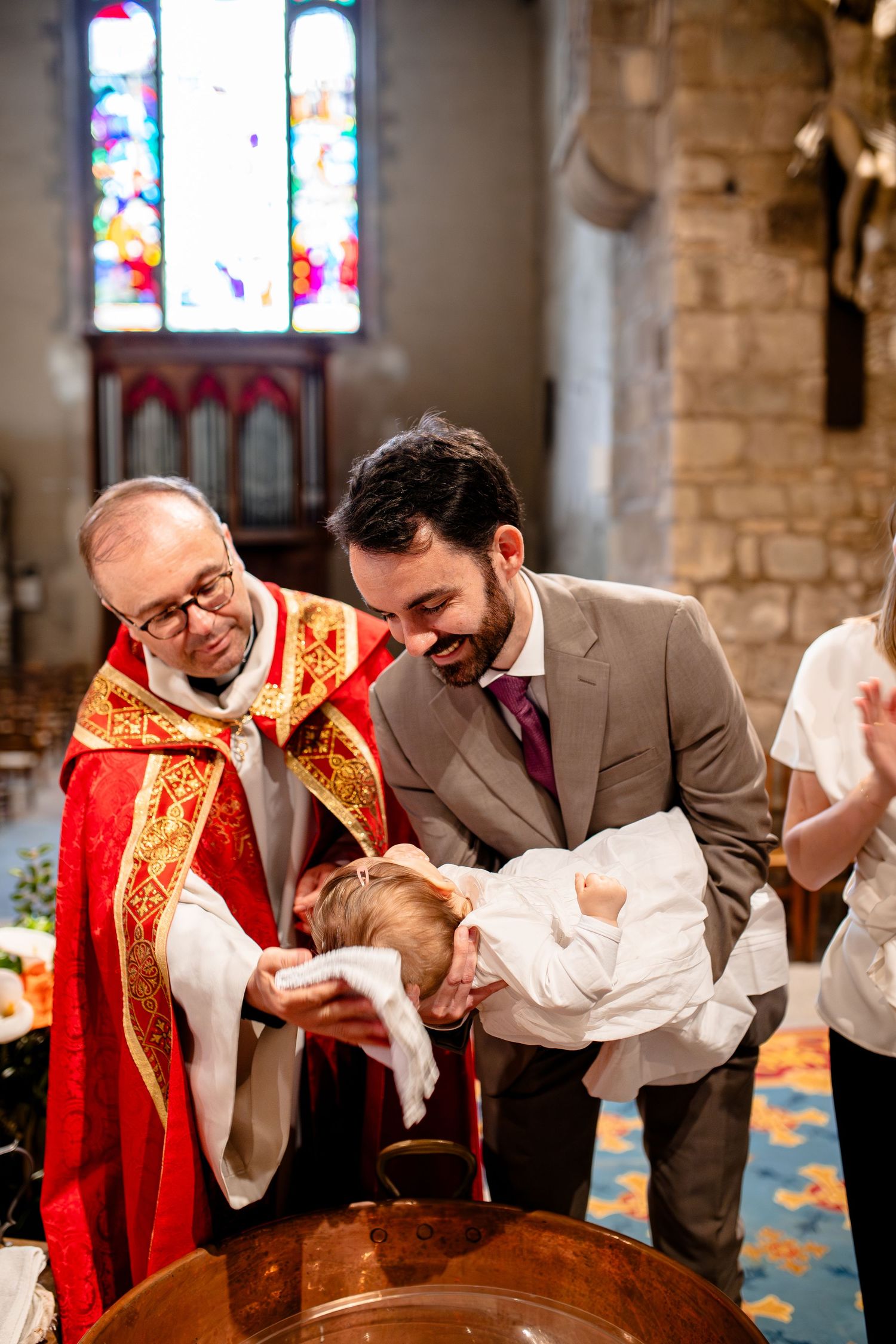 Clergy member performs a baptism ceremony inside a traditional stone church with stained glass windows.