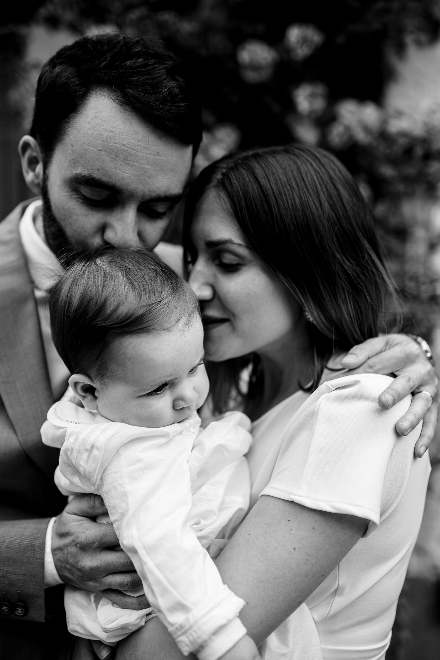 A tender black and white portrait of parents sharing a loving moment with their baby in white clothing.