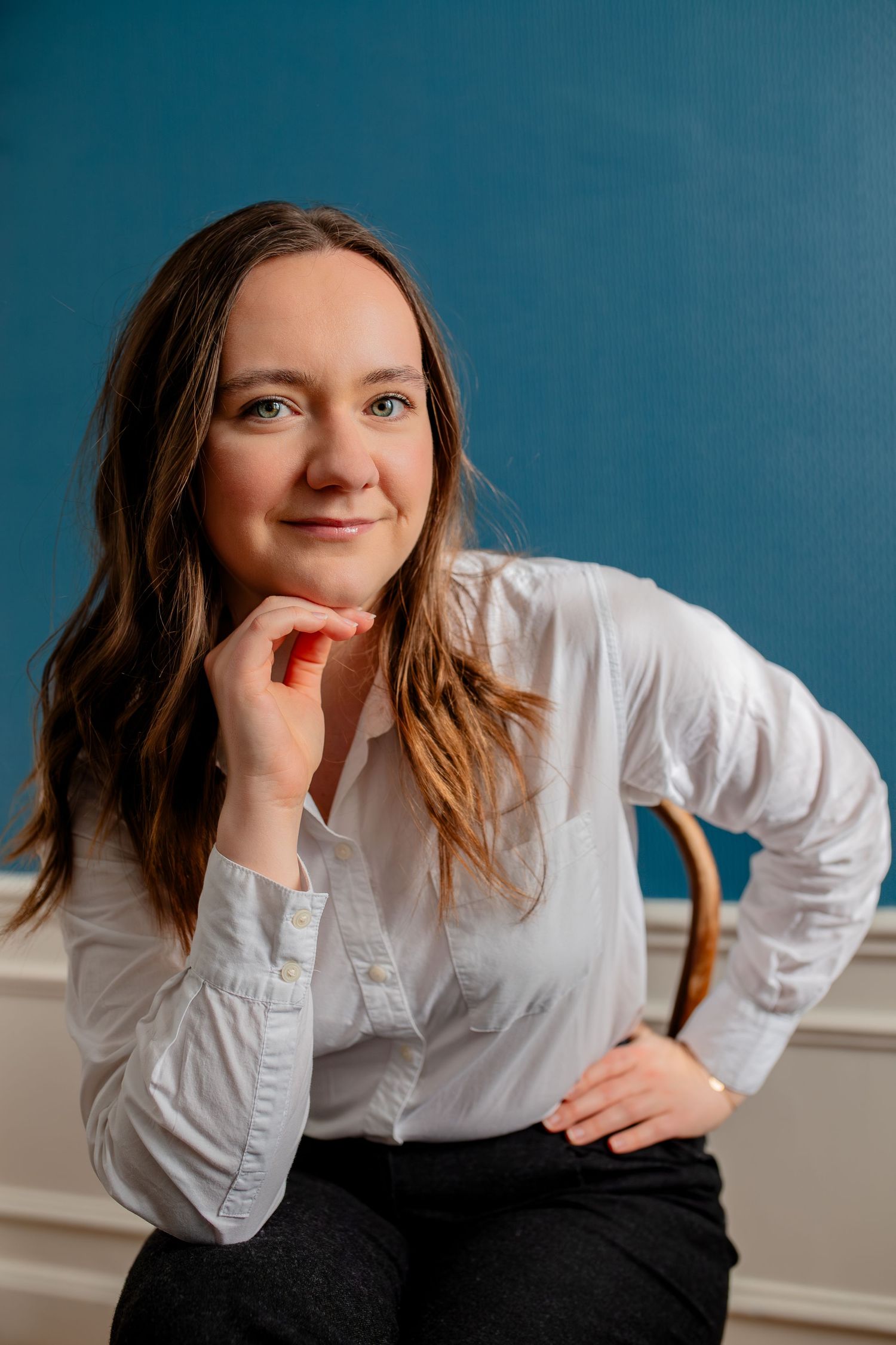 Professional headshot in white button-up shirt with hand on chin against blue background.