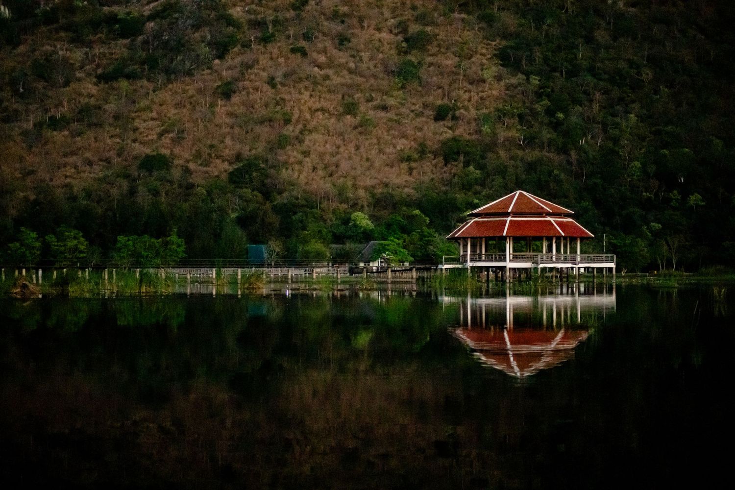 Red gazebo pavilion reflected in calm lake water against a forested hillside.