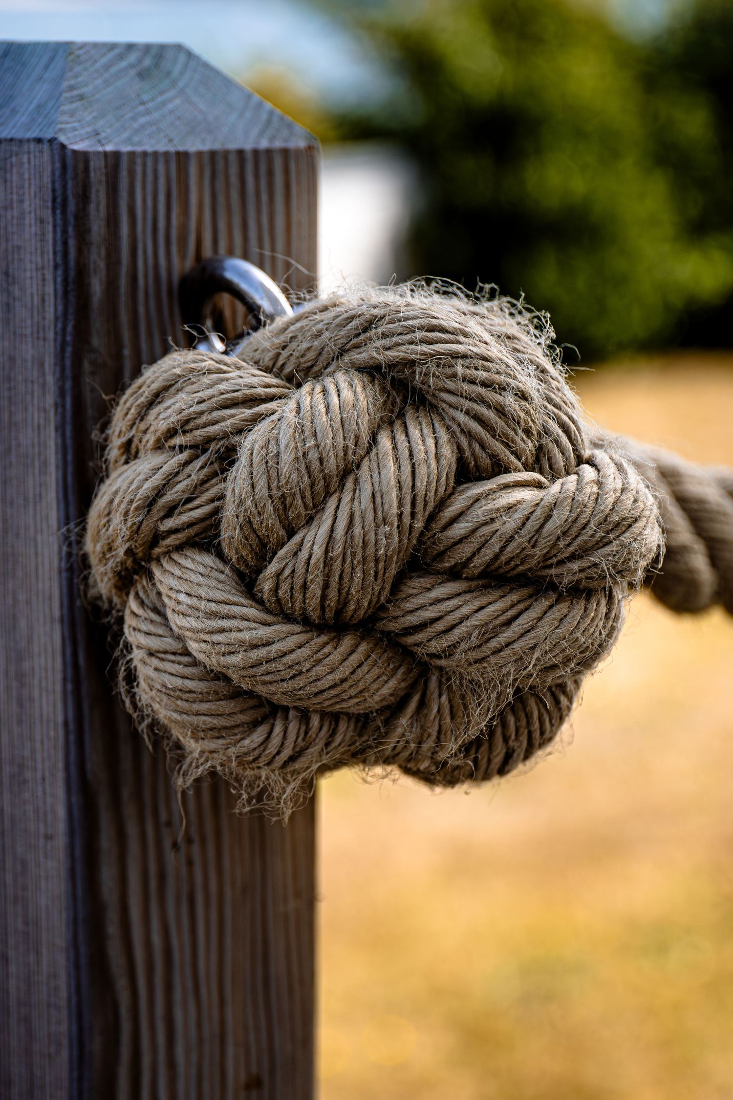 A braided rope knot made of thick beige natural fiber secured to a wooden post in outdoor setting.