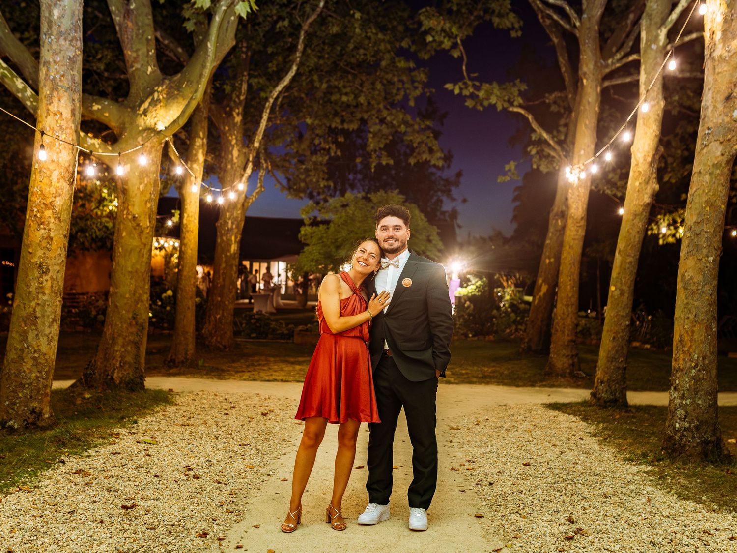 A couple stands on a gravel path at night with string lights illuminating trees in a romantic outdoor setting.