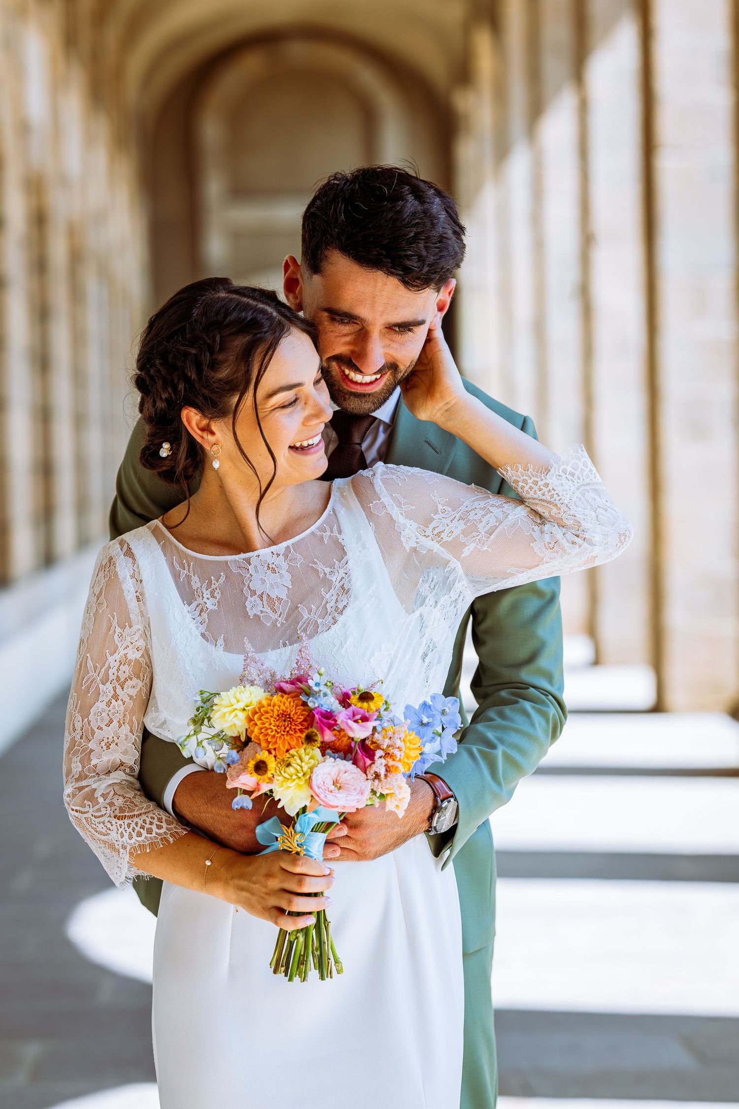 Newlywed couple embraces in a sunlit colonnade while holding a vibrant multicolored wedding bouquet.